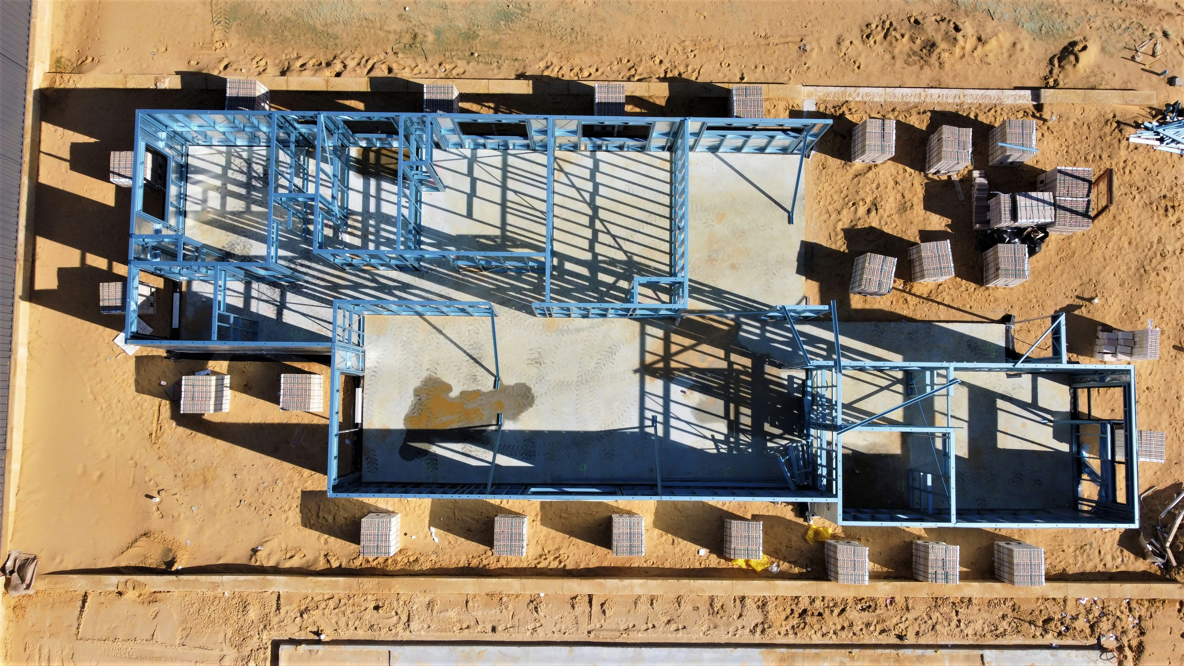 Aerial view of a house under construction showing metal framing on a concrete foundation surrounded by sand.