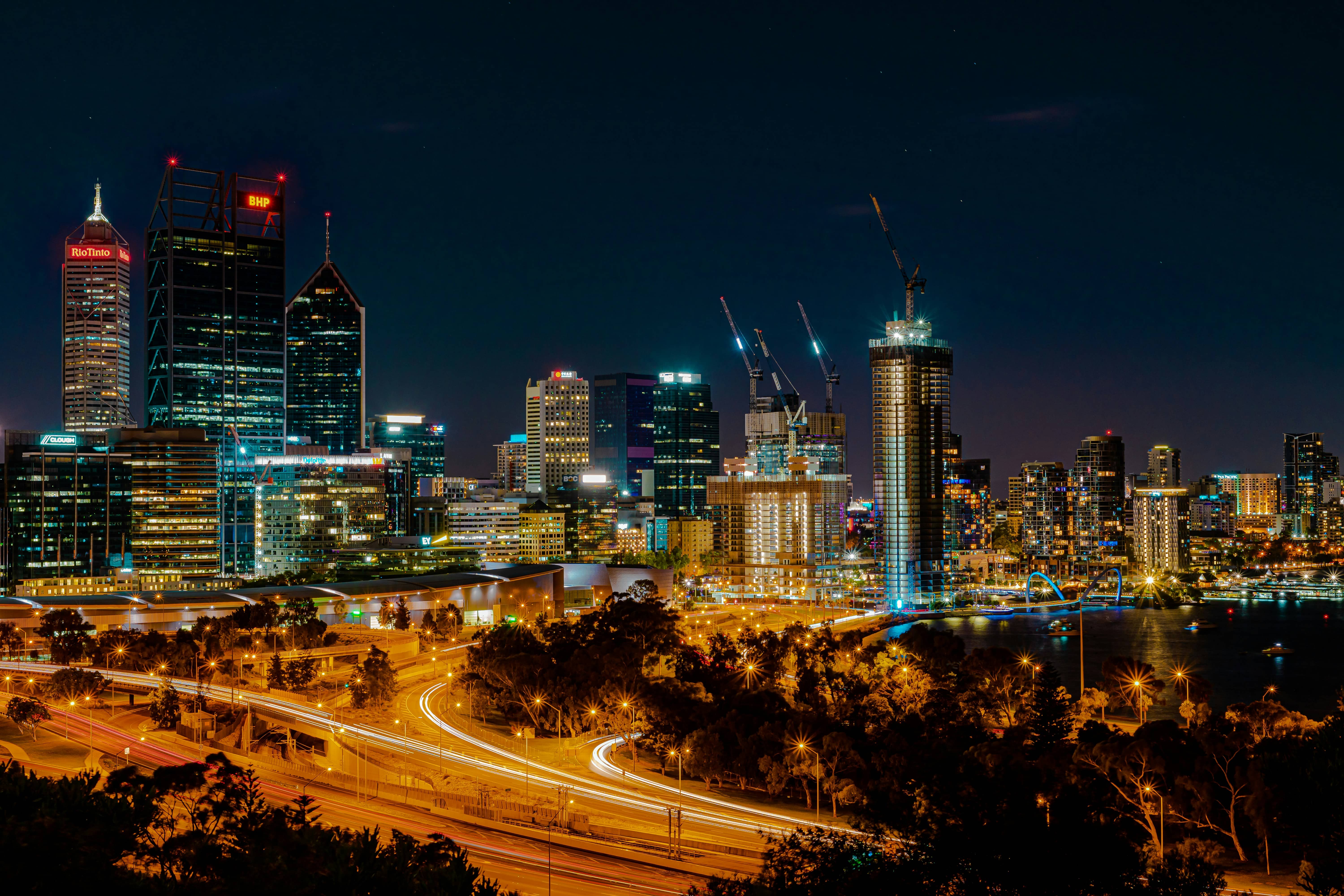 Night skyline of Perth city with illuminated skyscrapers, construction cranes, and light trails on roads.