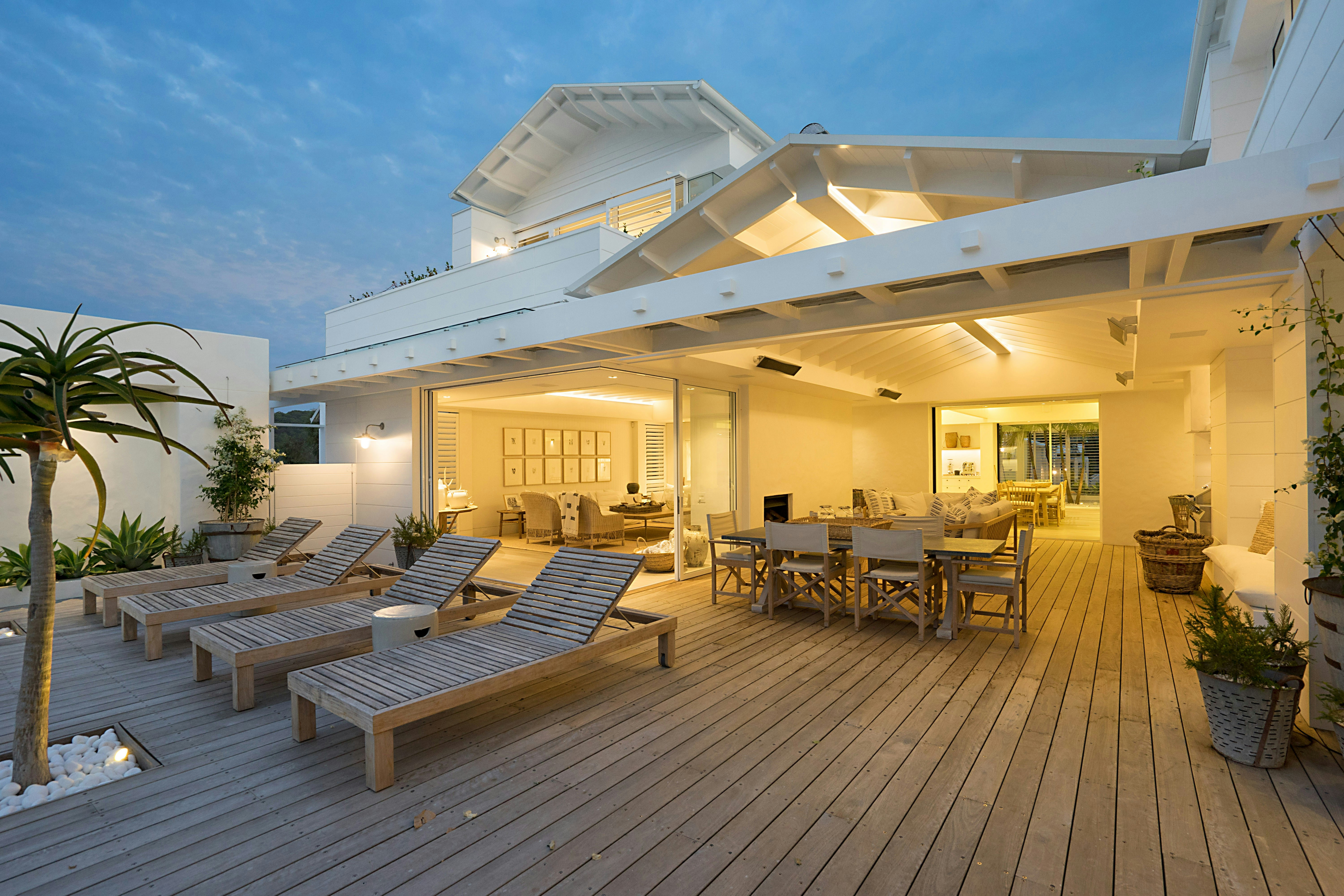 Spacious outdoor wooden deck with lounge chairs, dining table, and open living space of a modern white house at dusk.