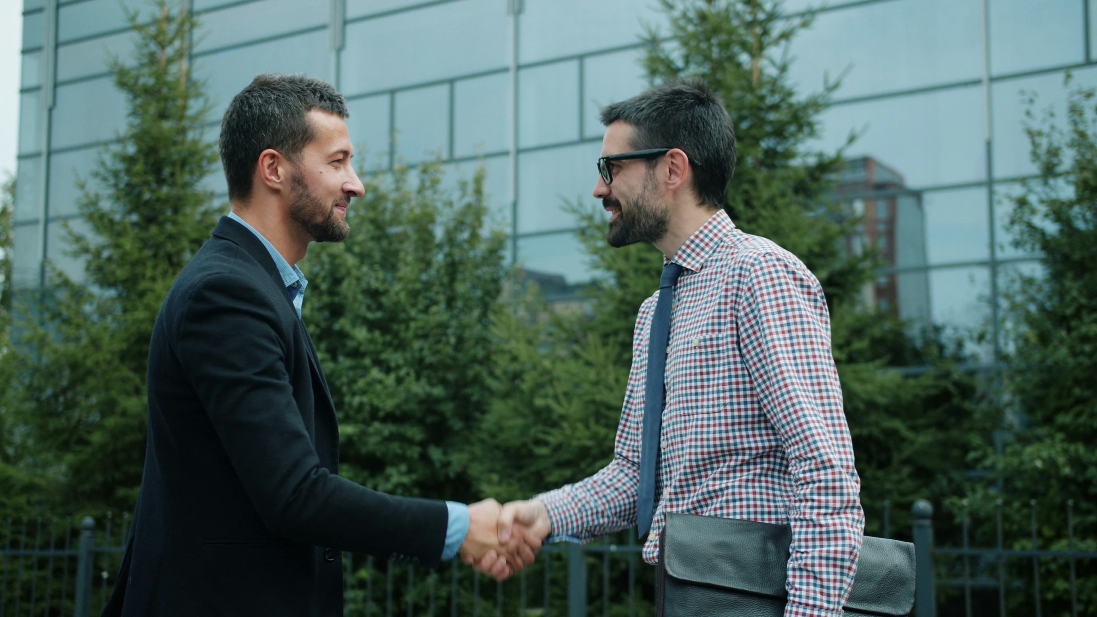Two men shaking hands outdoors in front of a building with glass windows and greenery.