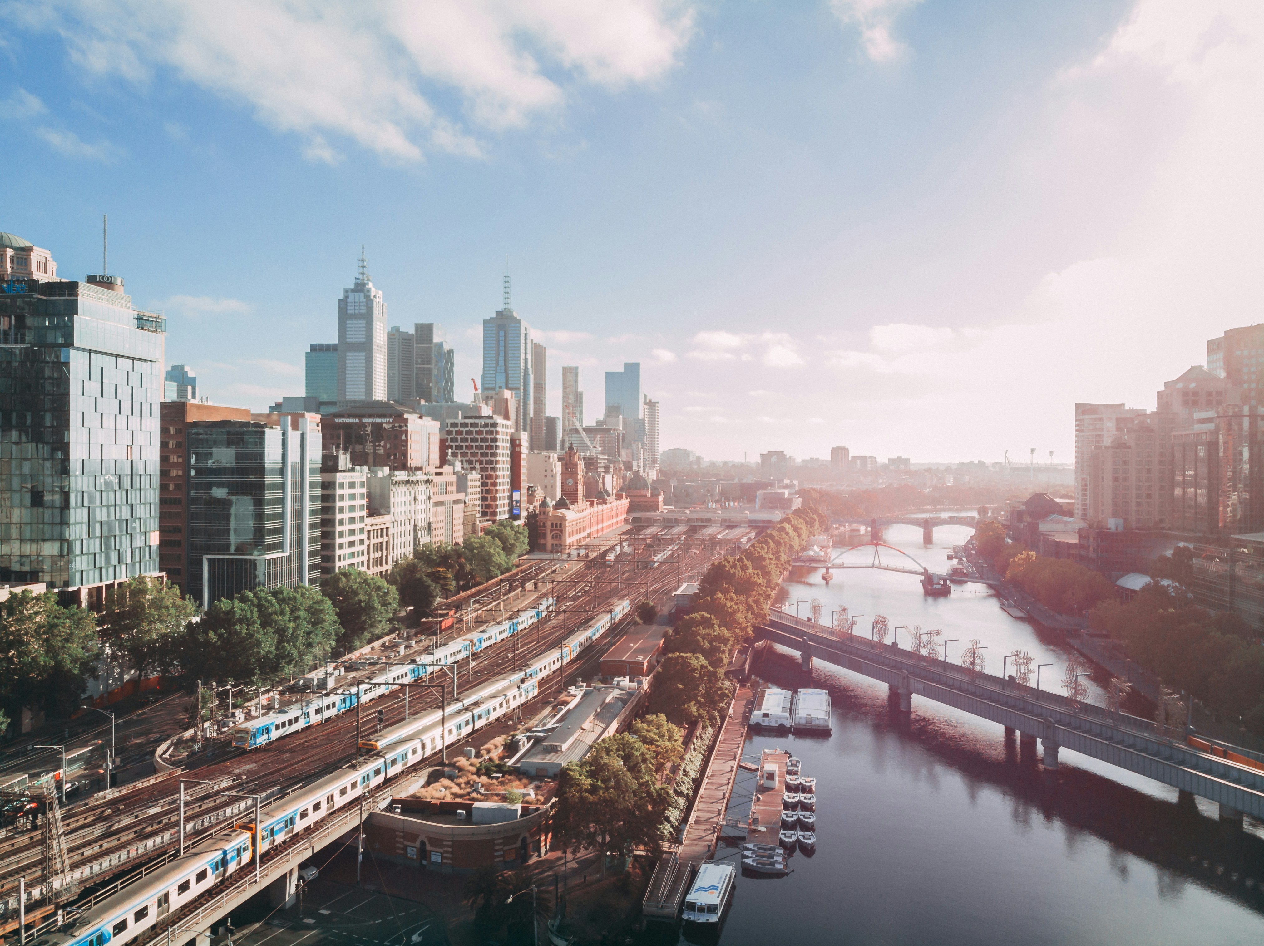 Cityscape of Melbourne showing railway tracks, trains, buildings, and a river with bridges under a partly cloudy sky.
