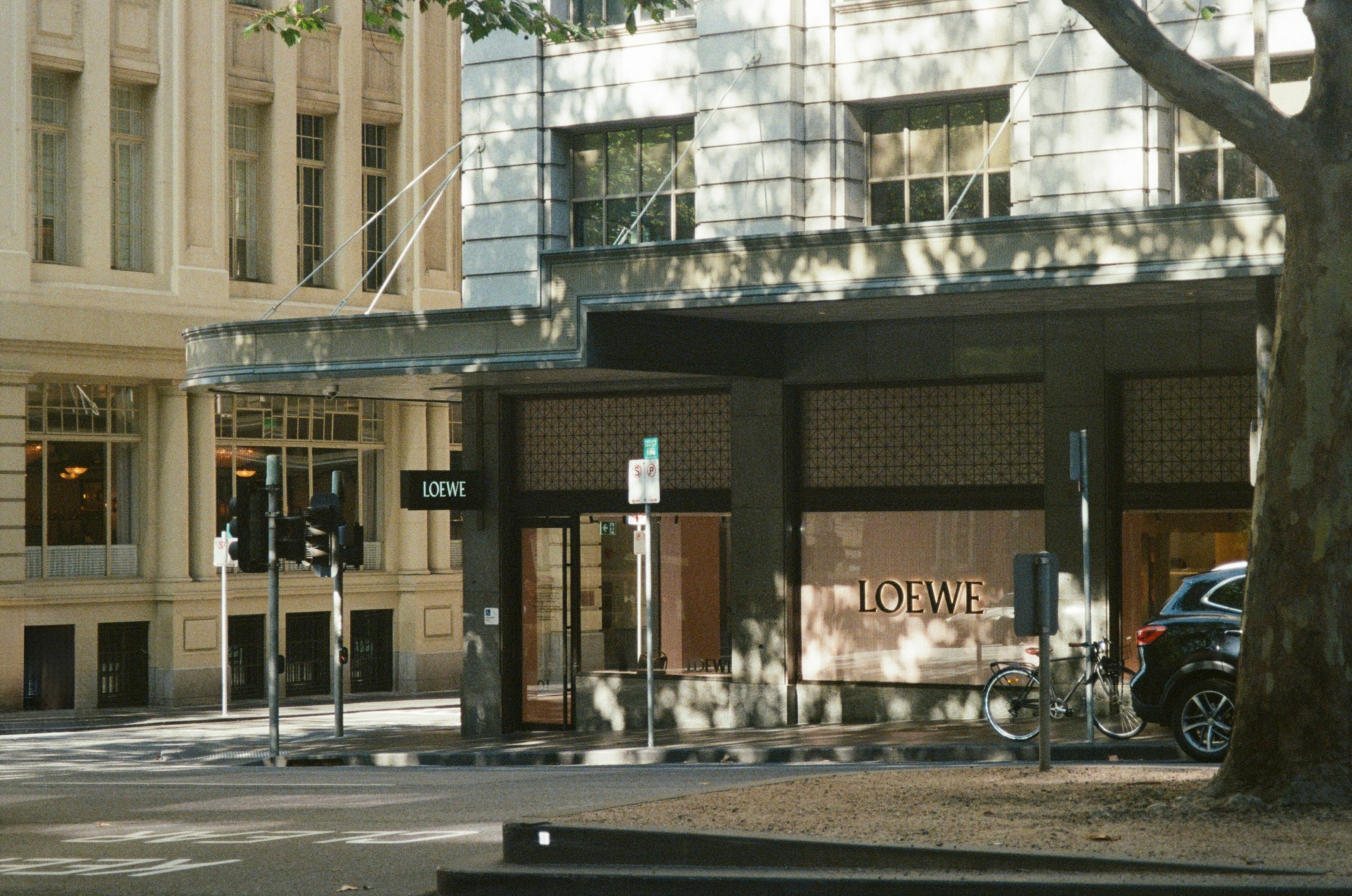 Exterior of Loewe luxury store on a street corner with trees casting shadows.