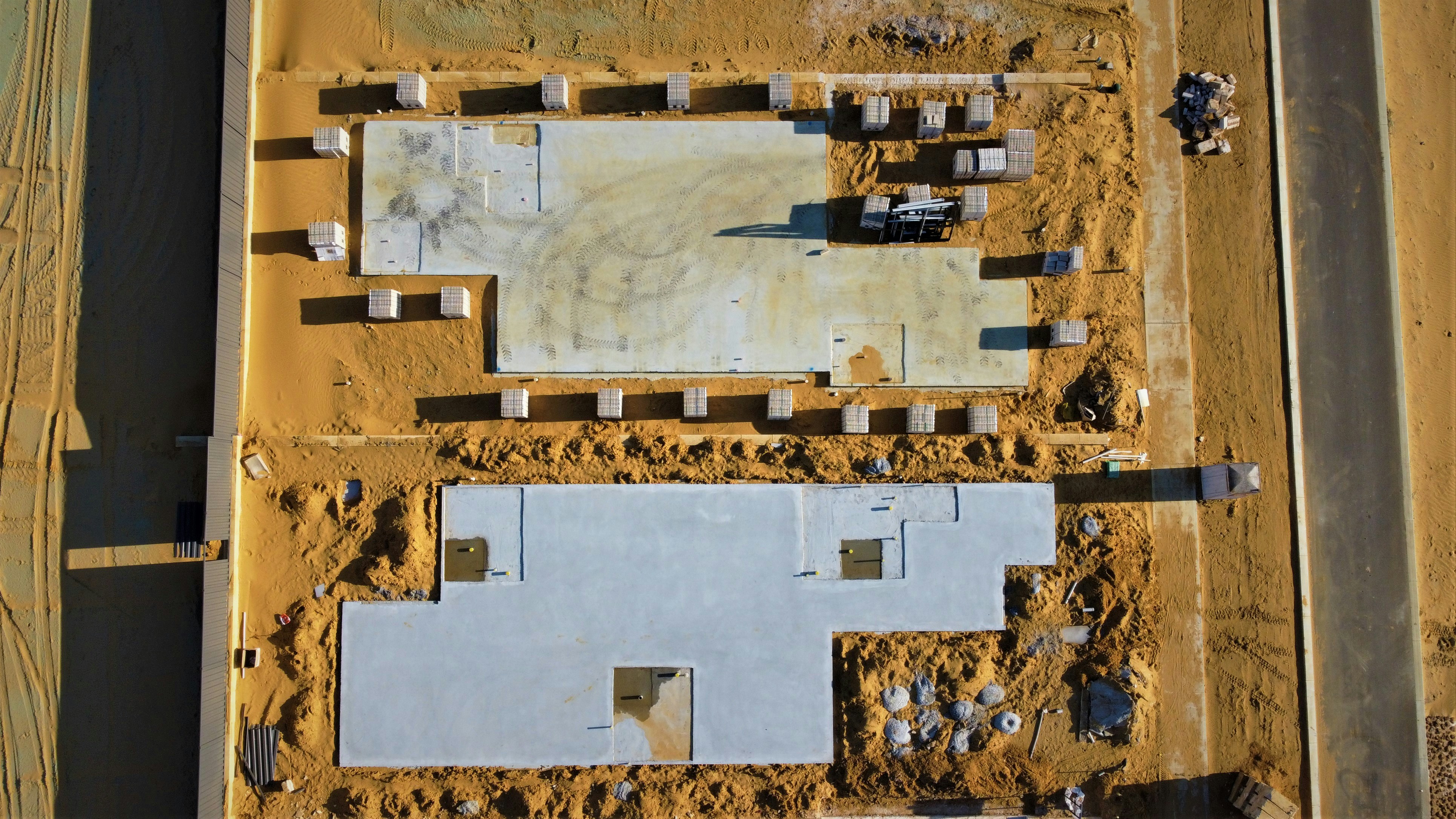 Aerial view of two concrete house foundations in a sandy construction site next to a paved road.