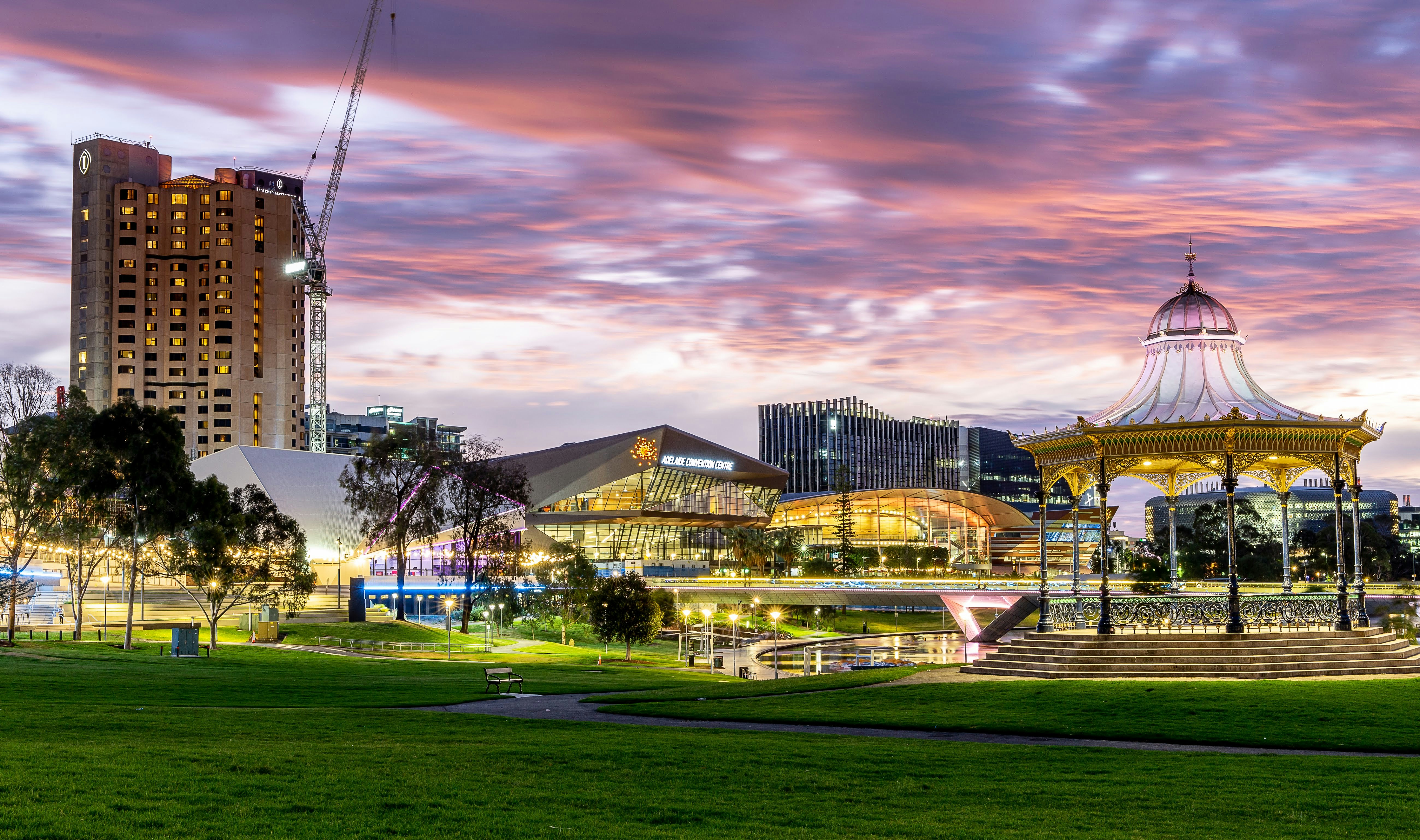 Adelaide cityscape at dusk with illuminated bandstand, green park, and modern convention center under a purple and pink cloudy sky.