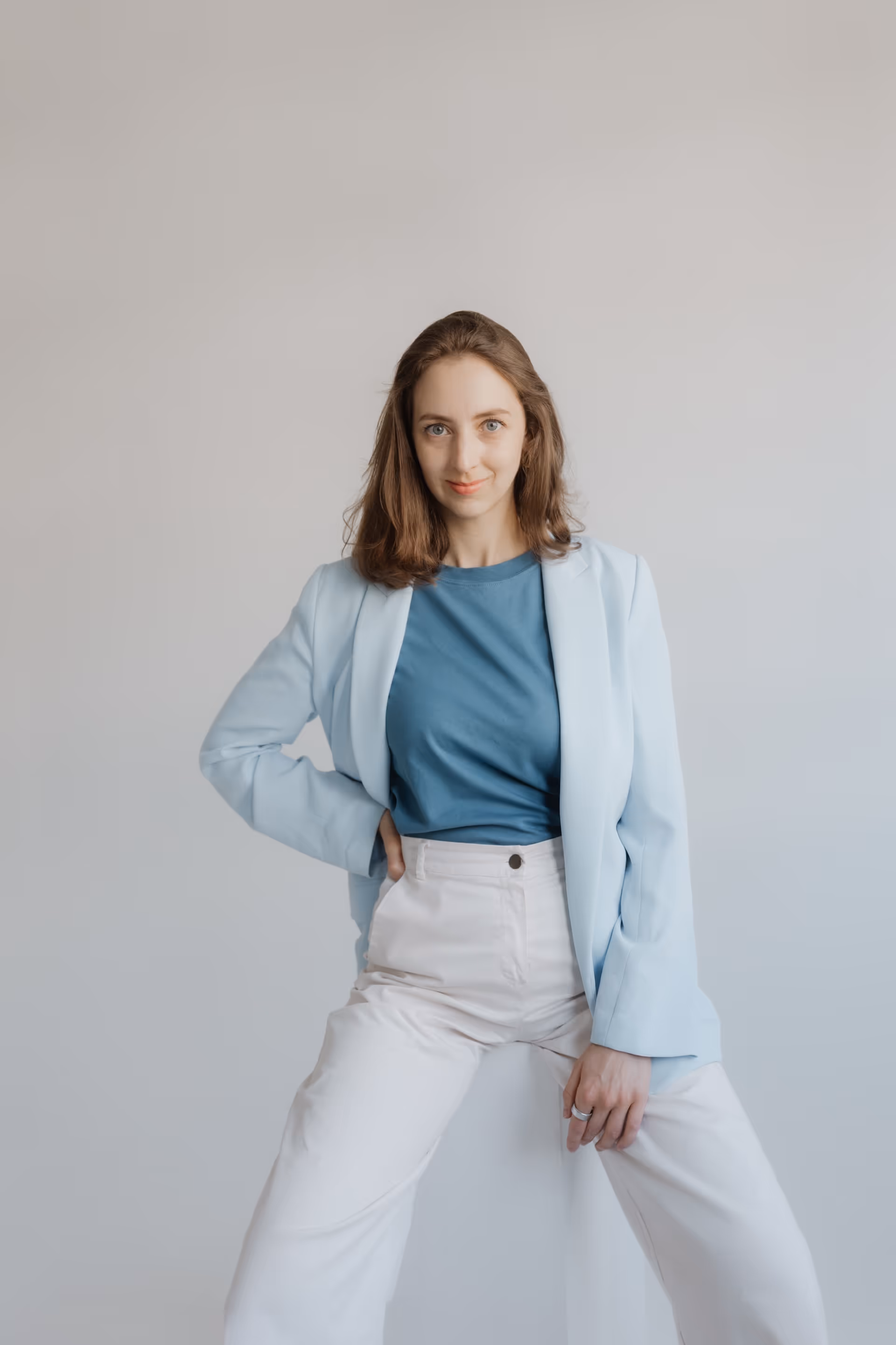 Natalie Ecker in blauem Blazer und weißer Hose sitzt vor grauem Hintergrund, lächelt leicht.