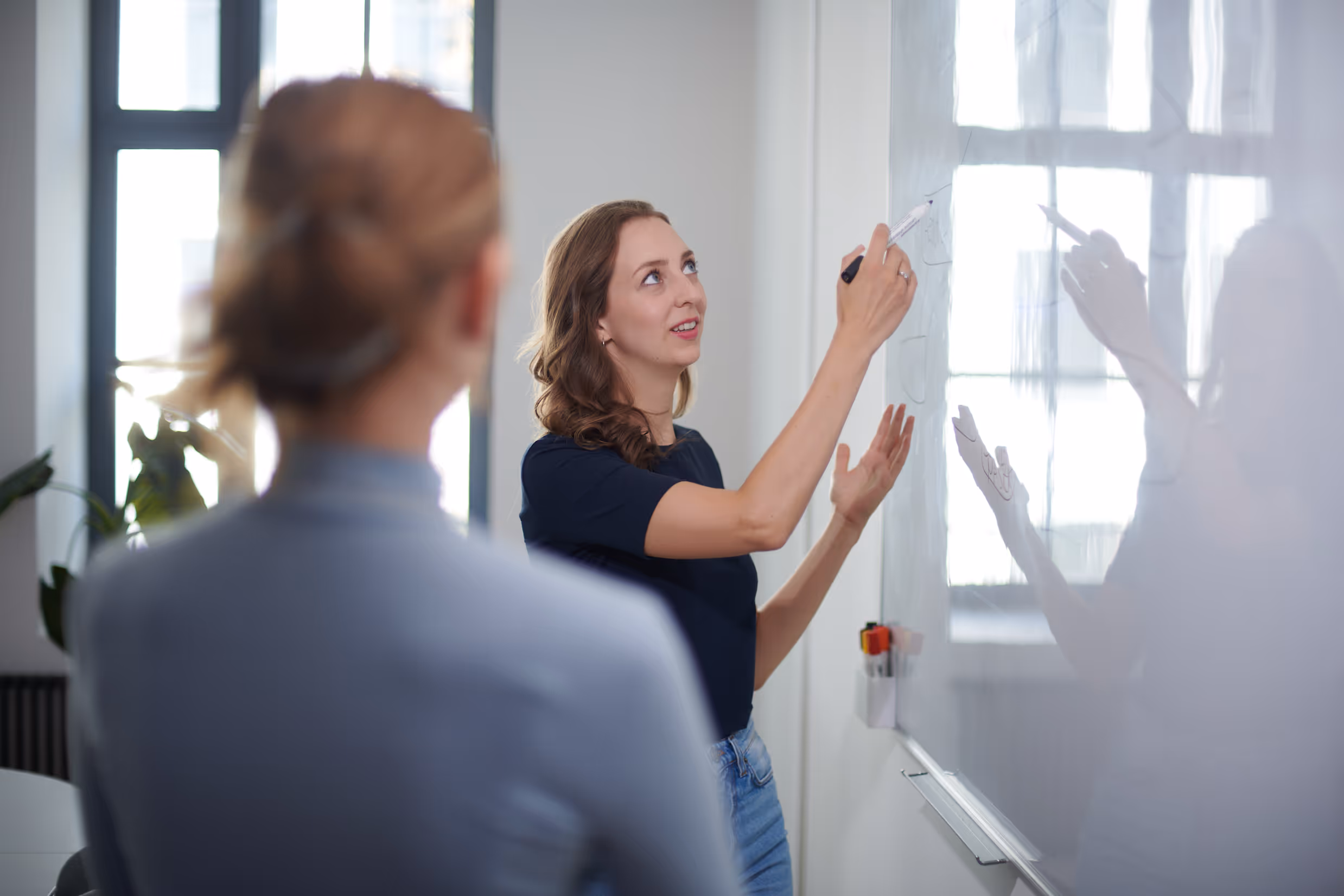 Natalie Ecker erklärt etwas mit Marker an Whiteboard, andere Person hört zu, in modernem Büro.