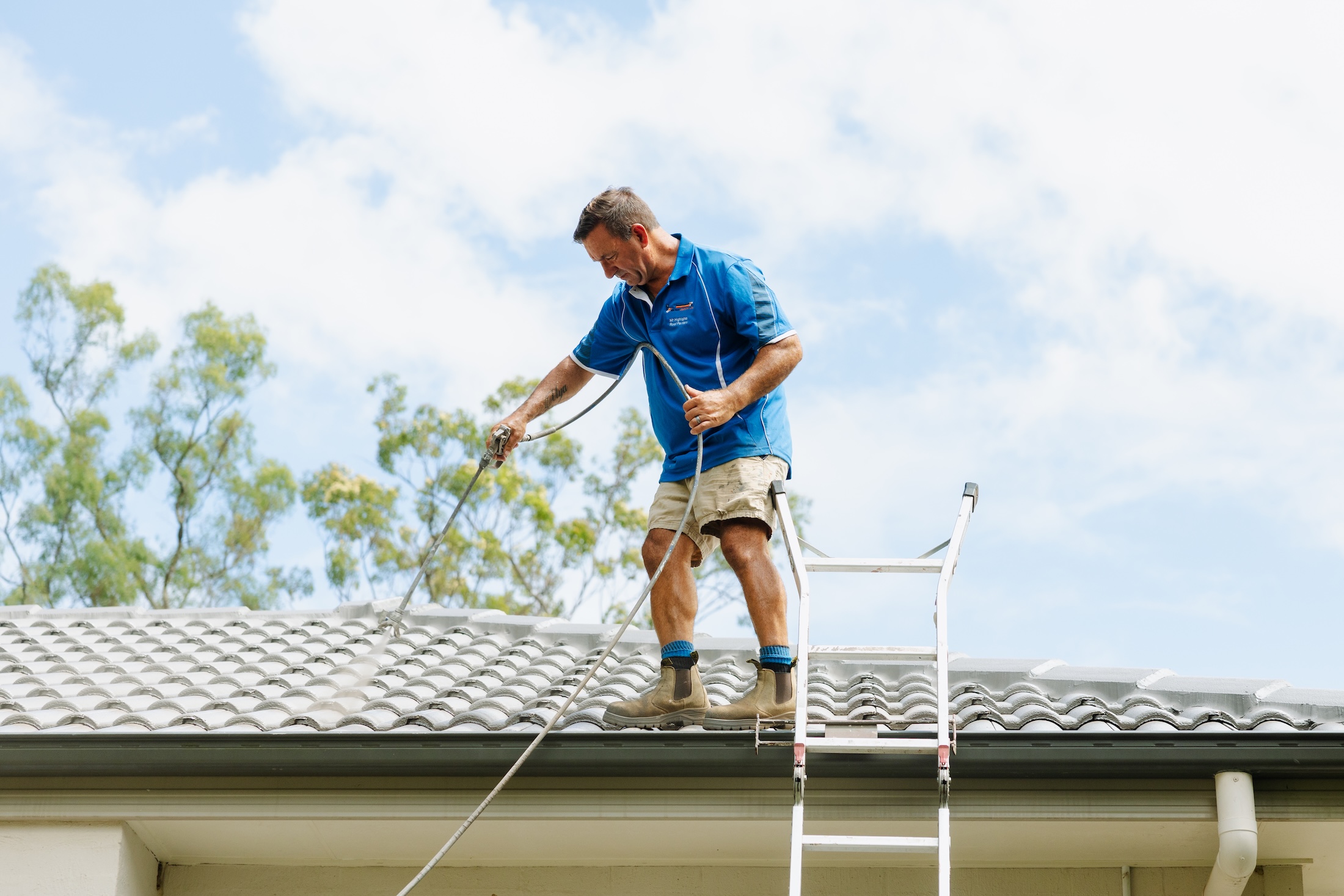 Mr Highlights Roofing team member using an airless sprayer to apply a heat reflective roof coating to a light grey concrete tile roof.