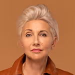 Portrait of a confident middle-aged woman with short blonde hair wearing a brown jacket against a brown background.