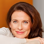 Smiling woman with brown hair resting her chin on her crossed arms against an orange background.