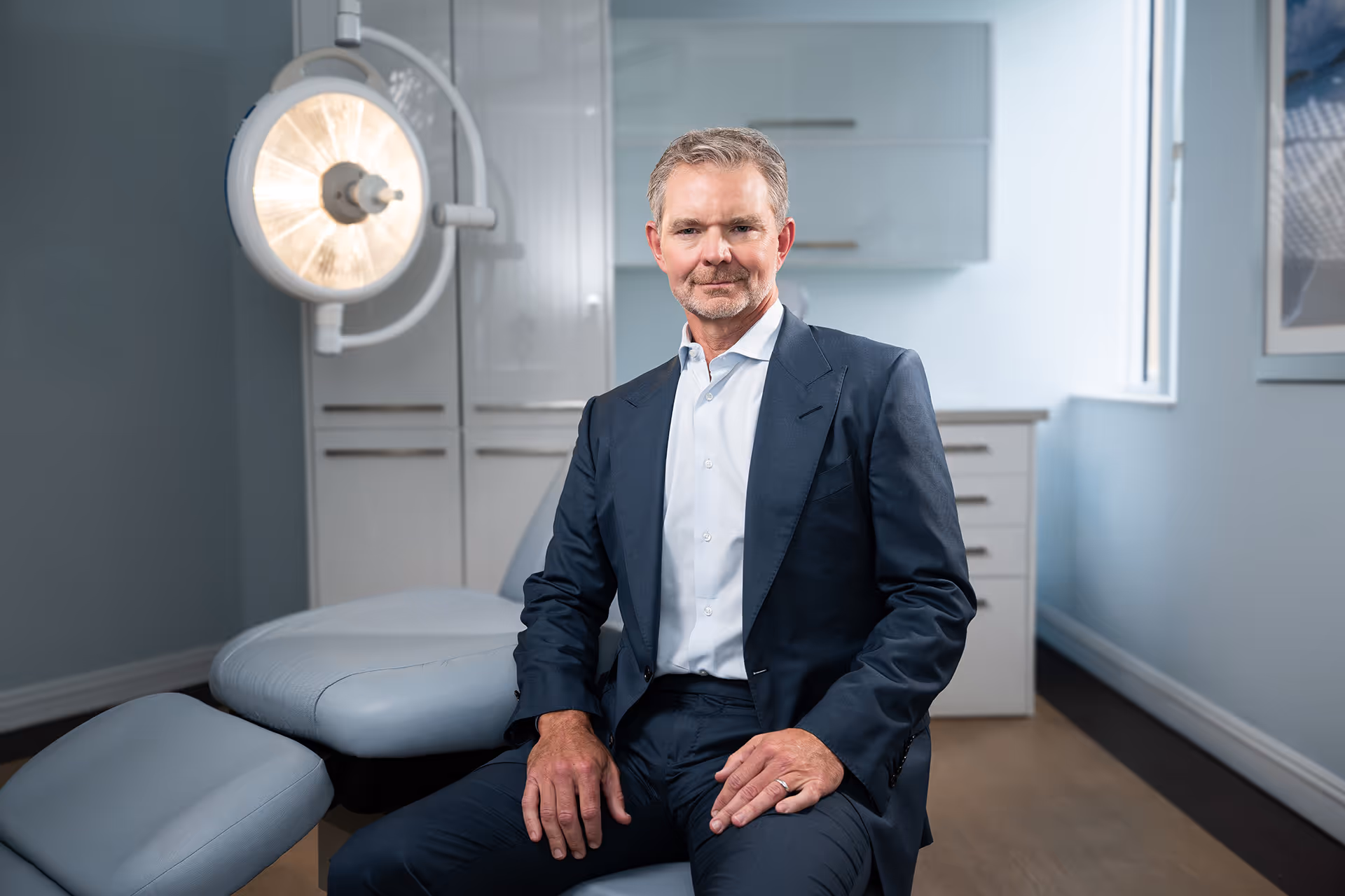 Middle-aged man in a dark suit sitting in a modern, clean medical examination room with a lighted surgical lamp behind him.
