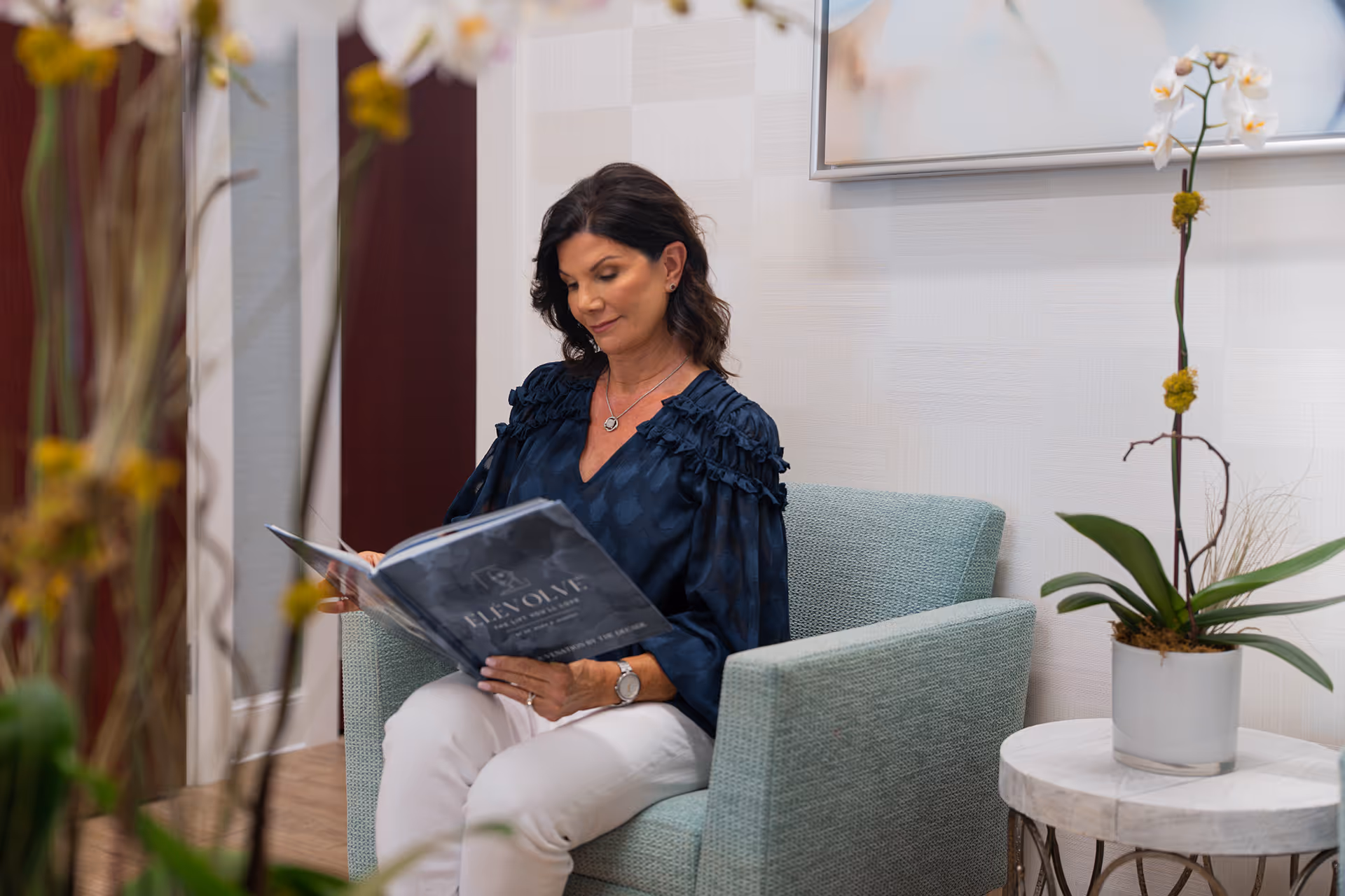 Woman sitting on light blue armchair reading a book titled 'Evolve' in a modern room with orchid on a side table.