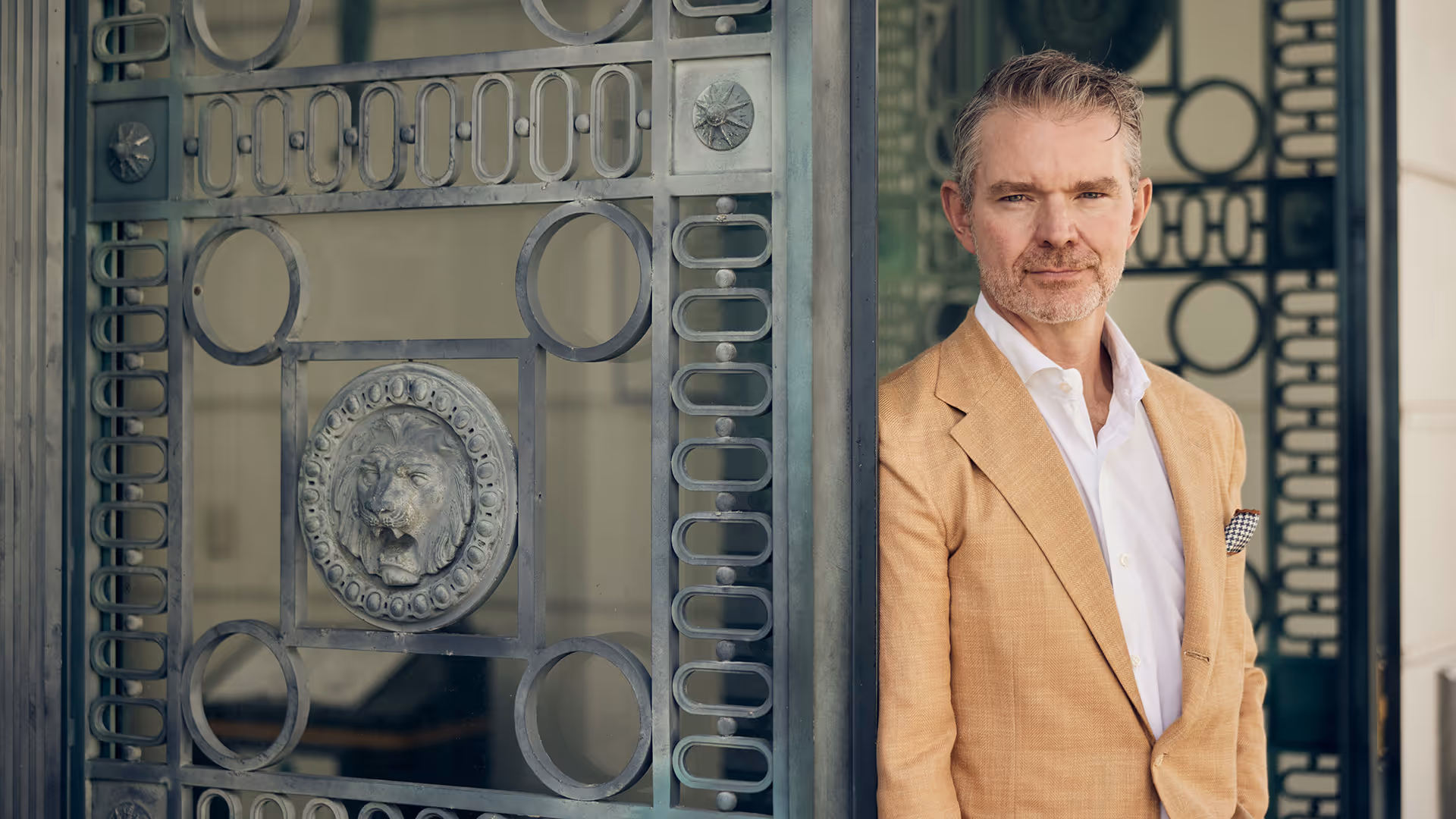 Middle-aged man in a tan blazer and white shirt standing beside an ornate metal gate with a lion head decoration.