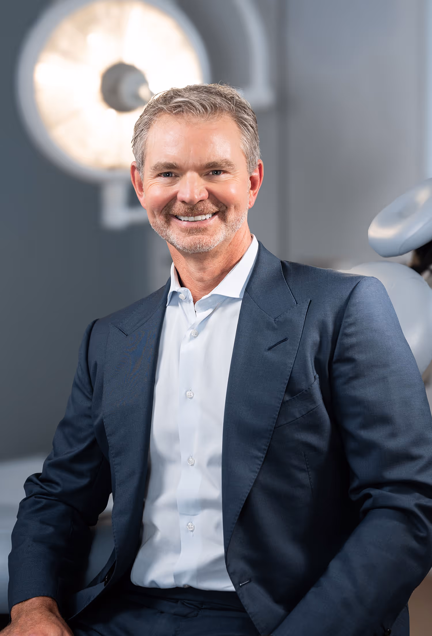 Smiling middle-aged man with gray hair and beard wearing a dark suit and white shirt, seated indoors with blurred background.