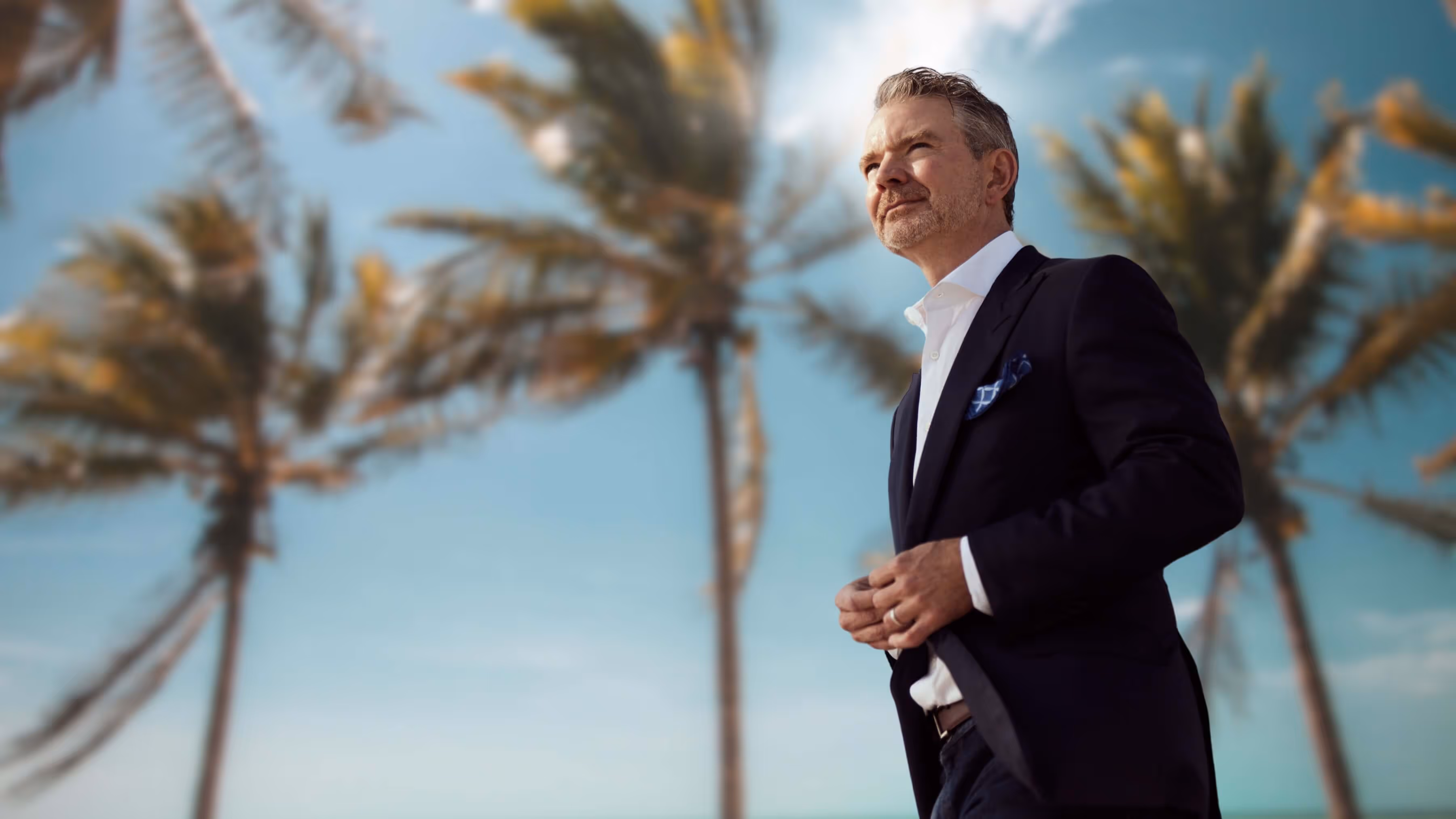 Man in a dark suit standing outdoors with blurred palm trees and blue sky in the background.