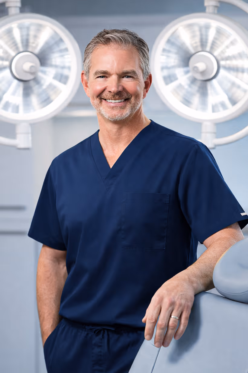 Smiling middle-aged male doctor in navy blue scrubs standing in a medical setting with surgical lights in the background.