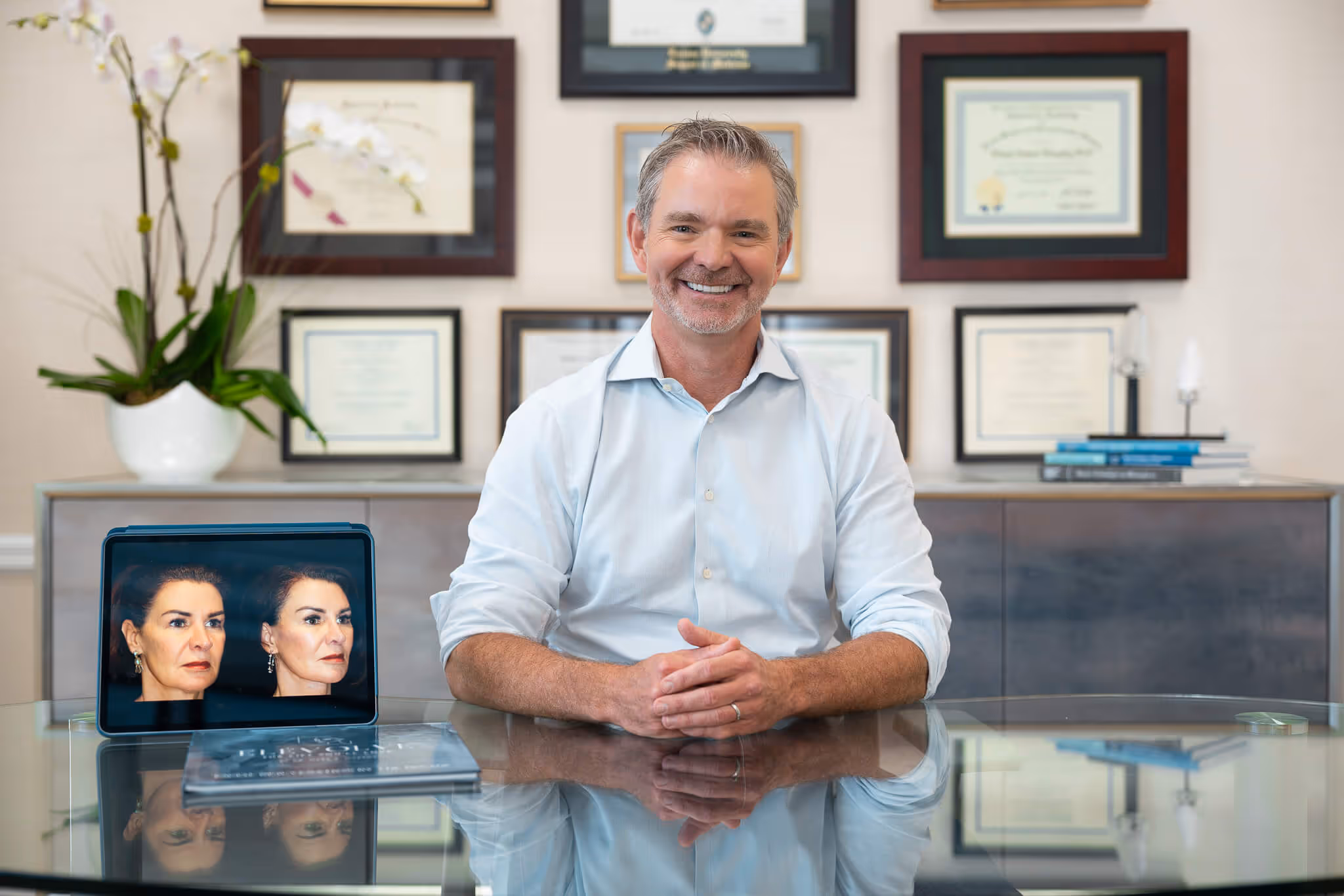 Smiling middle-aged man in a light blue shirt sitting at a glass table, with framed certificates on the wall behind him and a tablet displaying before and after facial photos.