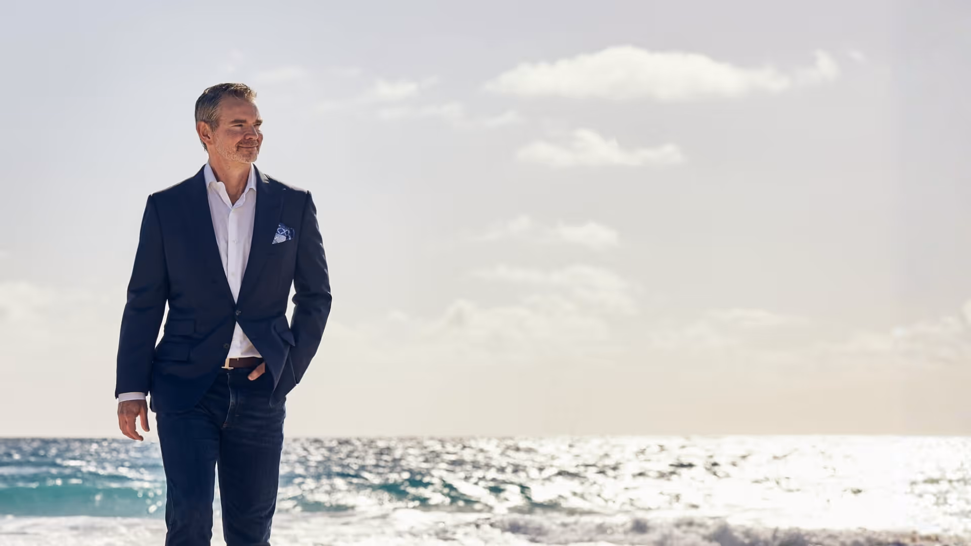 Man in a navy blazer and white shirt walking on a sunny beach with ocean waves in the background.