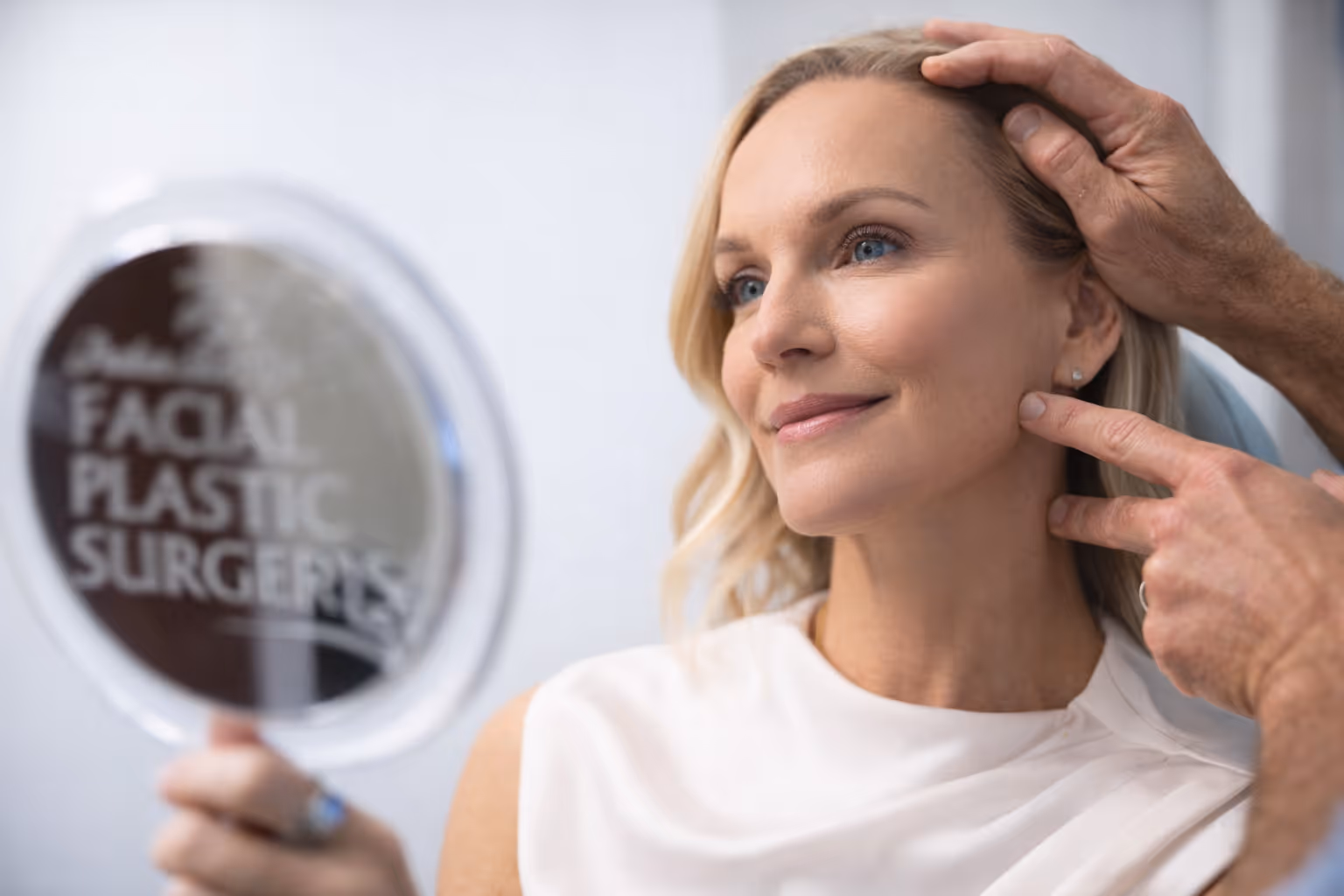Woman with blonde hair looking at her face in a handheld mirror while a man’s hands gently examine her jaw and cheek.