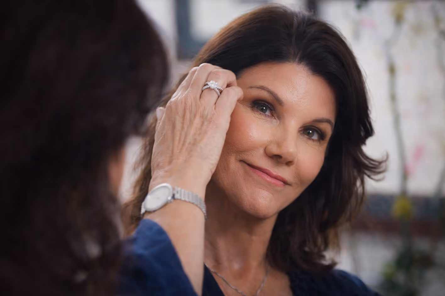 Middle-aged woman with dark hair gently touching her face and smiling.