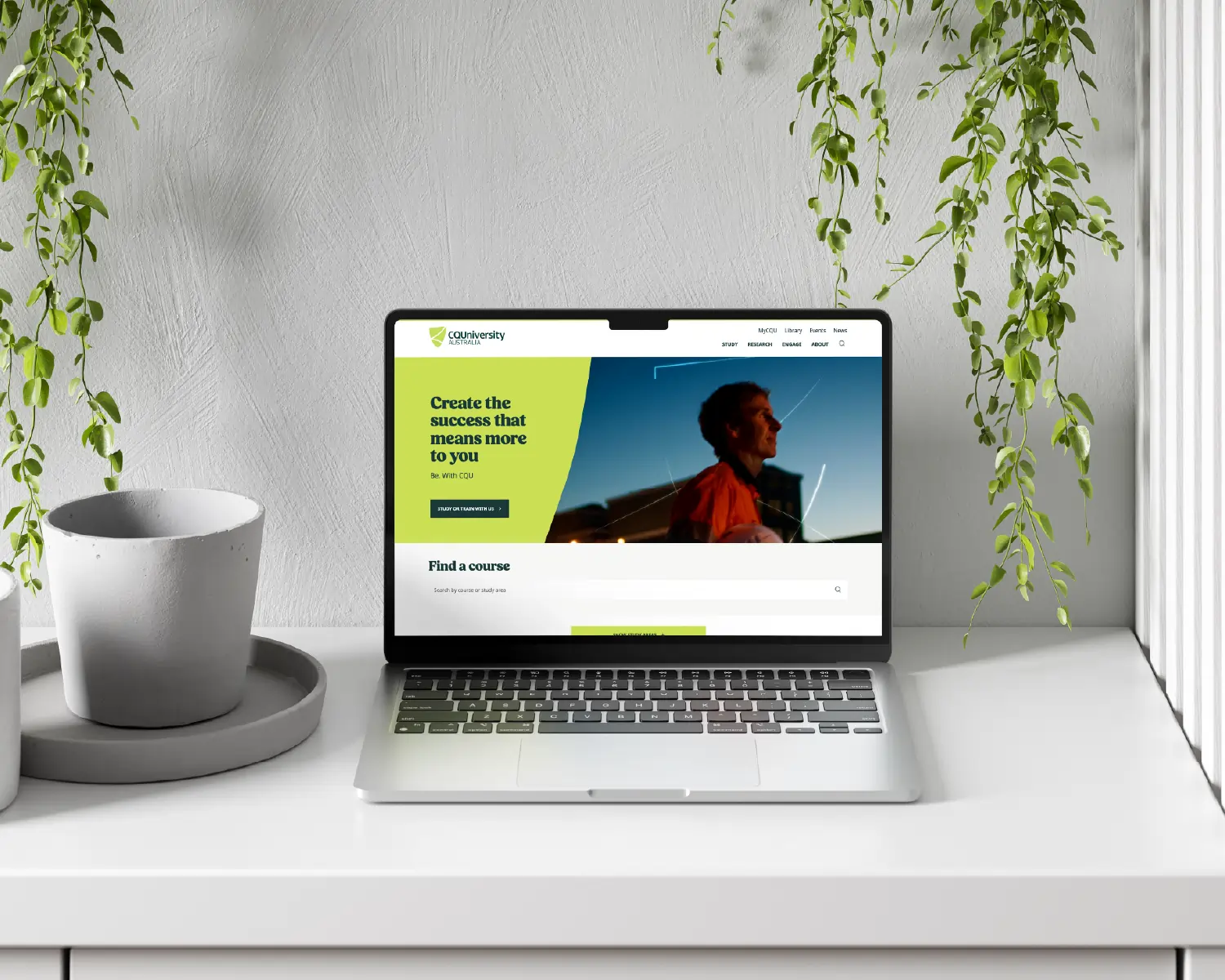 Laptop on white desk showing CQU University website with green text, next to empty pot and hanging plants.