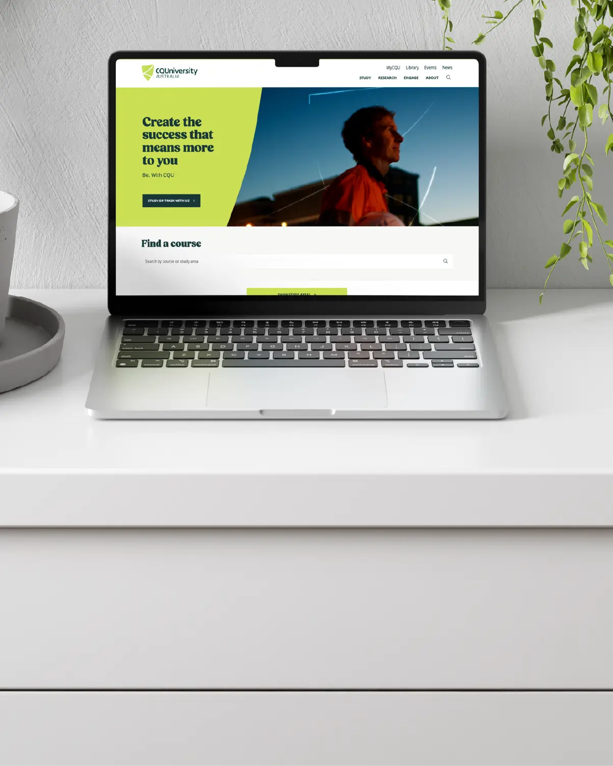 Laptop on white desk displaying CQUniversity website with a man in an orange jacket on screen.