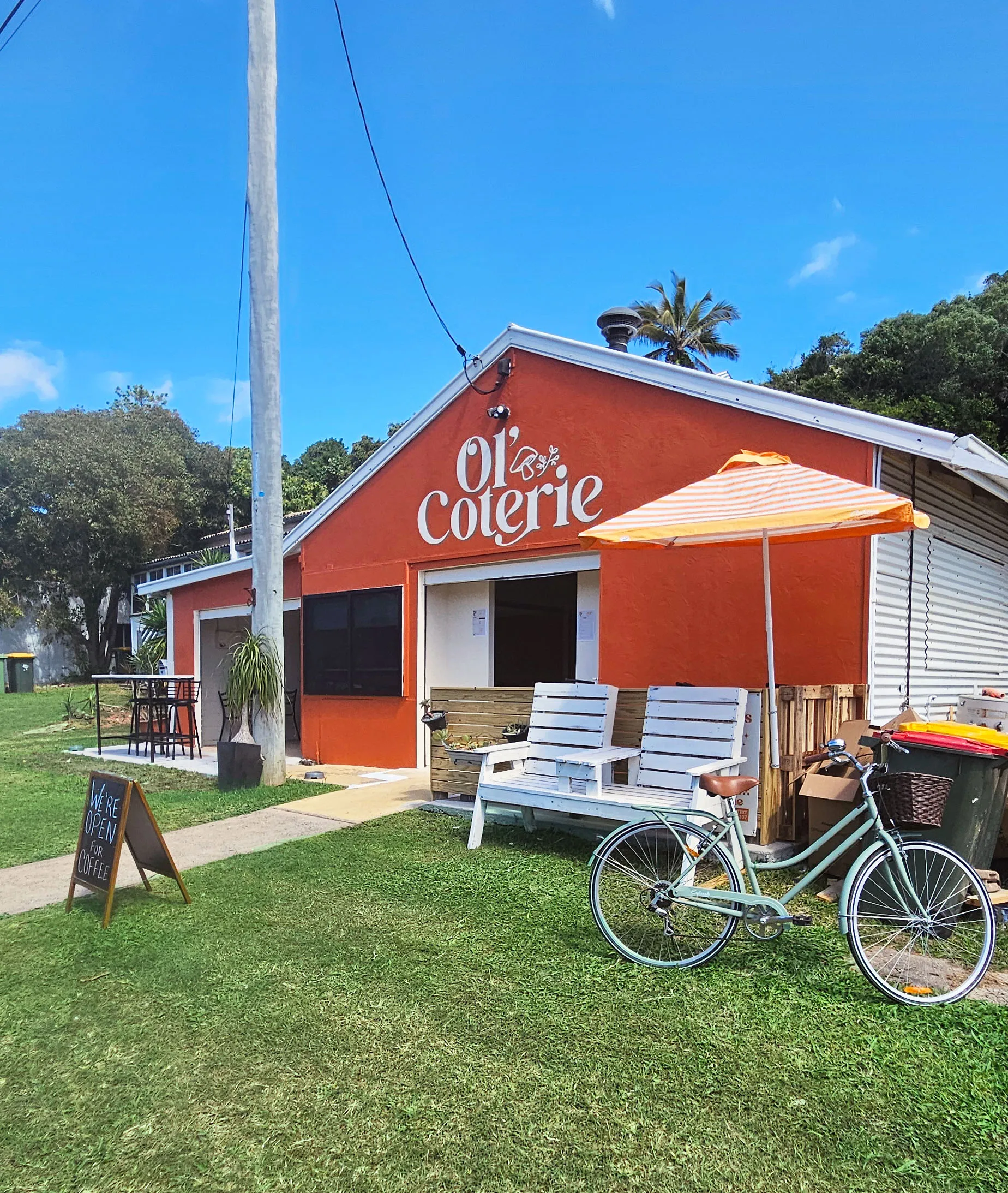 Orange building with 'ol' coterie' sign, white bench under striped umbrella, bicycle, and grassy area.