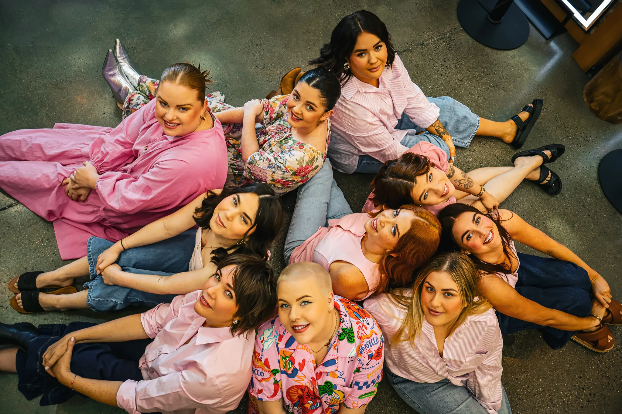 Group of nine women seated on the floor in a circle, smiling and looking up at the camera.