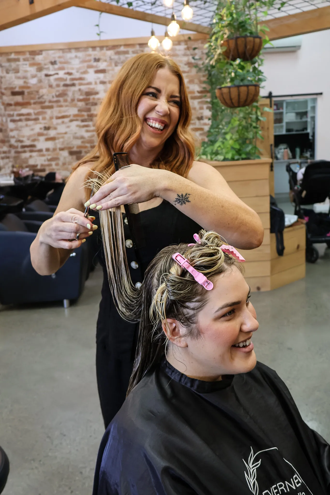 Smiling stylist cutting a seated woman's wet hair held up with pink clips in a modern salon.