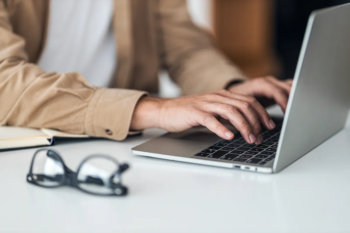 Hands typing on a silver laptop keyboard at a white desk with glasses and notebook nearby.