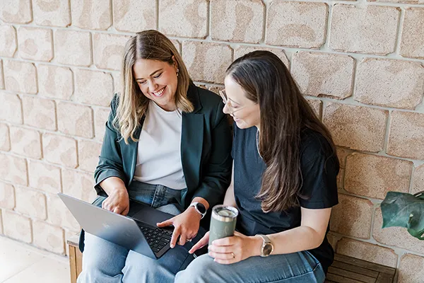 Two women sitting on a bench, smiling and looking at a laptop, one holding a tumbler cup.