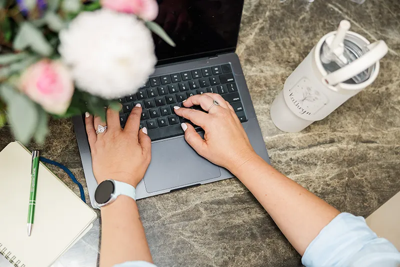Person typing on a laptop keyboard at a table with a notebook, pen, and a cup with straws.
