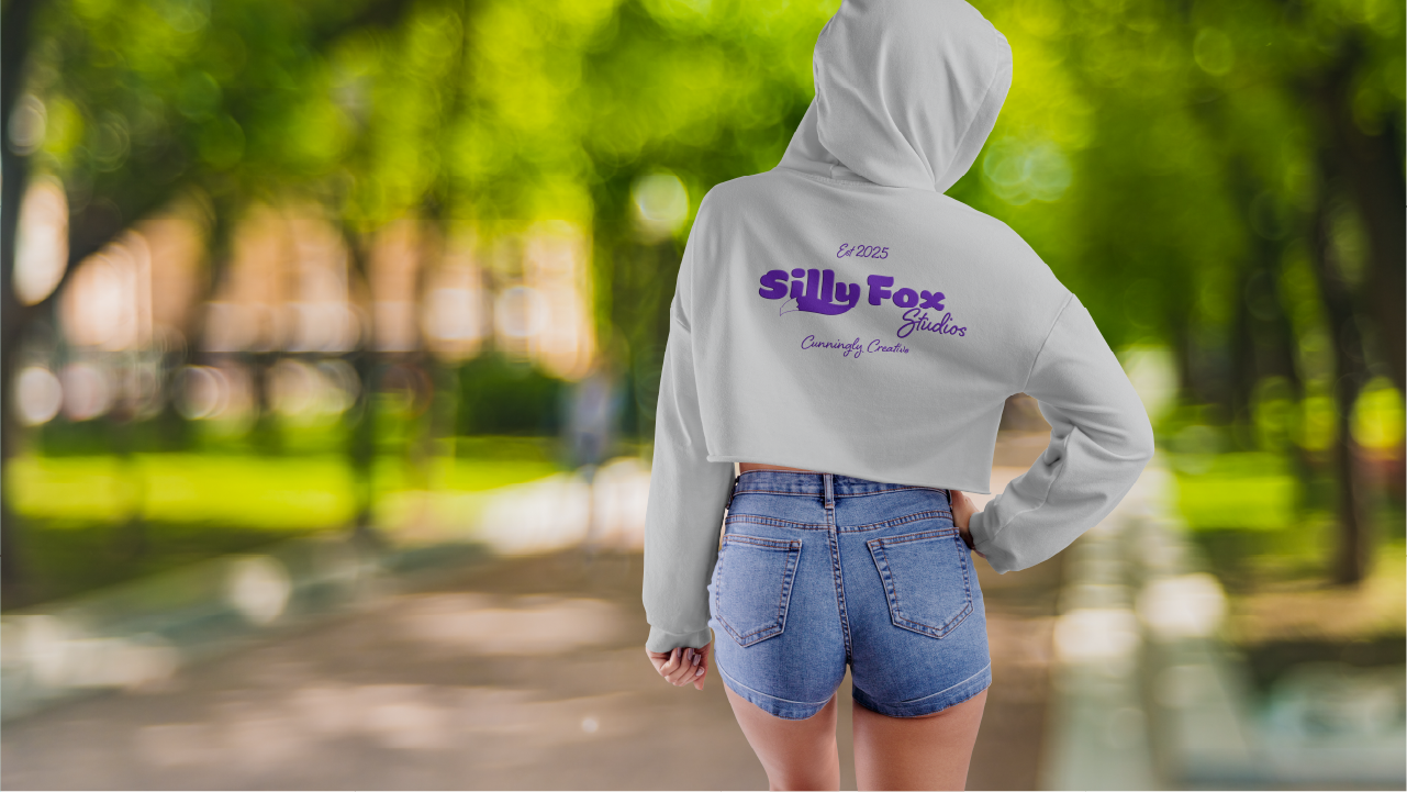 Person wearing a light gray cropped hoodie with the purple text "Silly Fox Studios Est 2025 Cunningly Creative" on the back, paired with blue denim shorts, standing on a blurred park pathway with green trees.