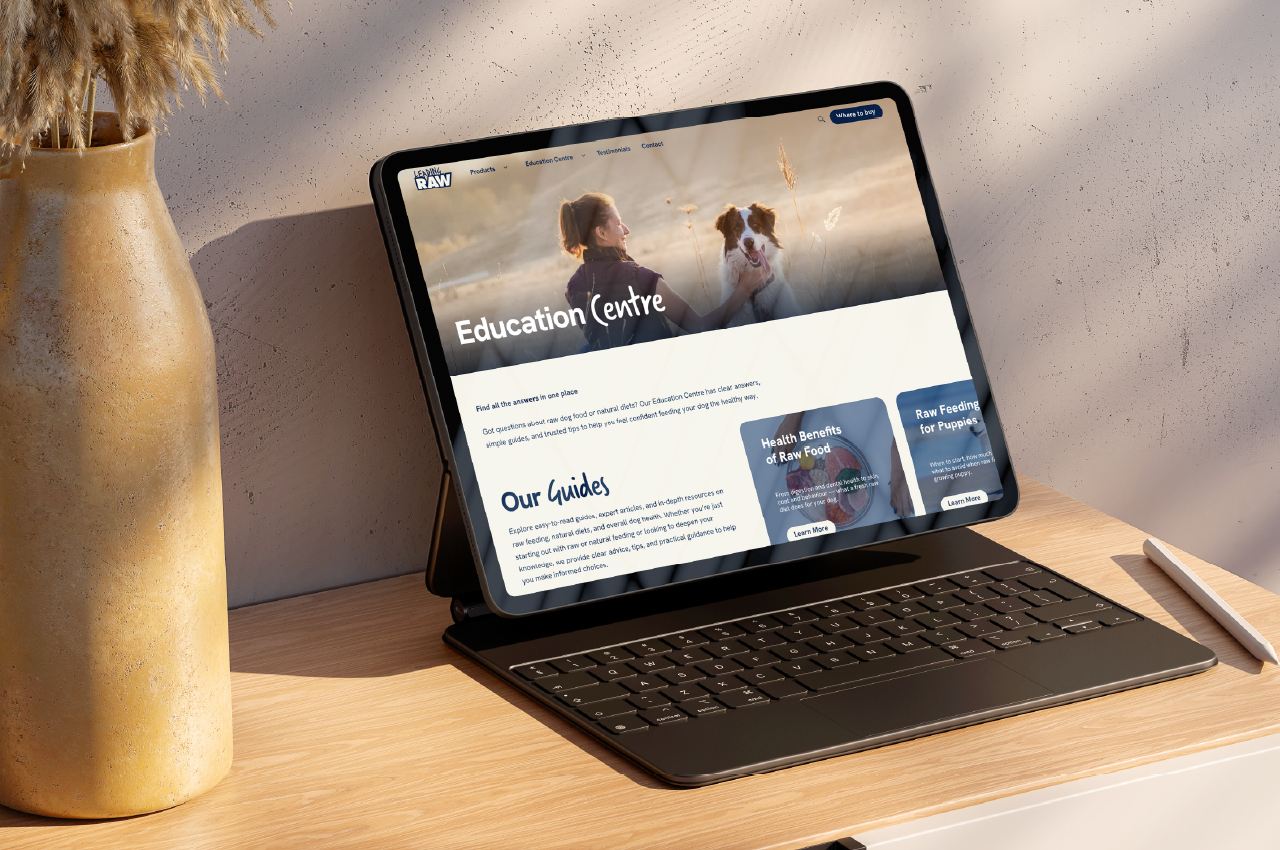 Tablet with keyboard on a wooden desk displaying a webpage titled Education Centre, featuring an image of a woman with a dog in a field, sections for guides on health benefits of raw food and feeding puppies, with a stylus pen beside it and a tall vase with dried plants in the background.