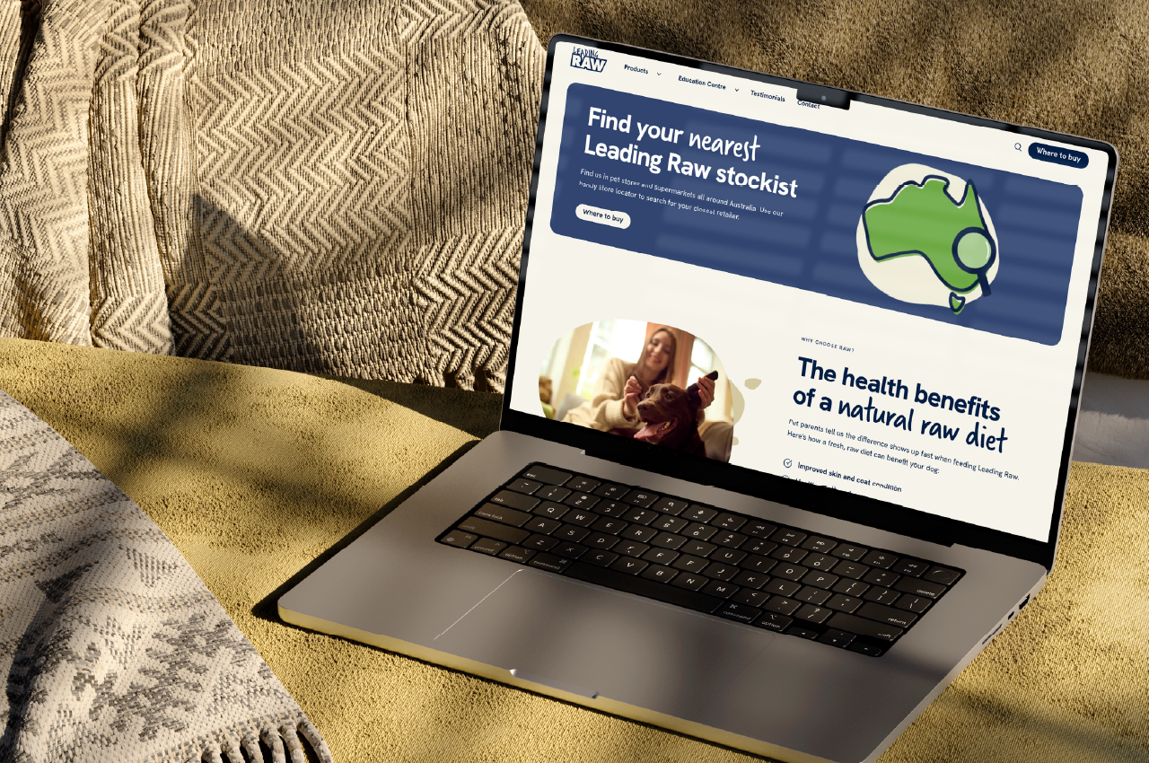 Open laptop on a textured yellow surface displaying a website with a blue banner stating 'Find your nearest Leading Raw stockist' and a section titled 'The health benefits of a natural raw diet' with an image of a woman with a dog.