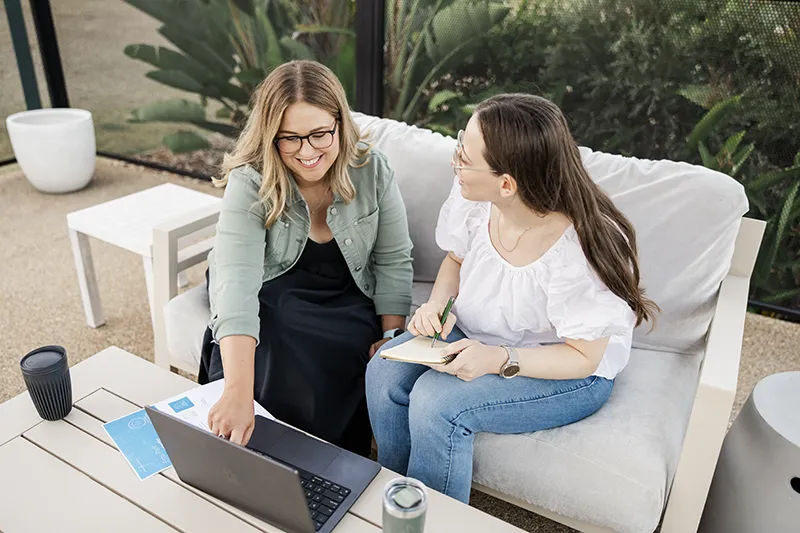 Two women sitting on a light gray sofa outside by a patio table, looking at a laptop together; one woman is pointing at the laptop screen while the other takes notes in a notebook.