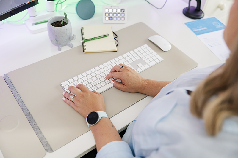 Person typing on a white keyboard at a desk with a mouse, notebook with a pen, small plant in a gray pot, a smartwatch on the left wrist, and a paper with text partially visible on the right.