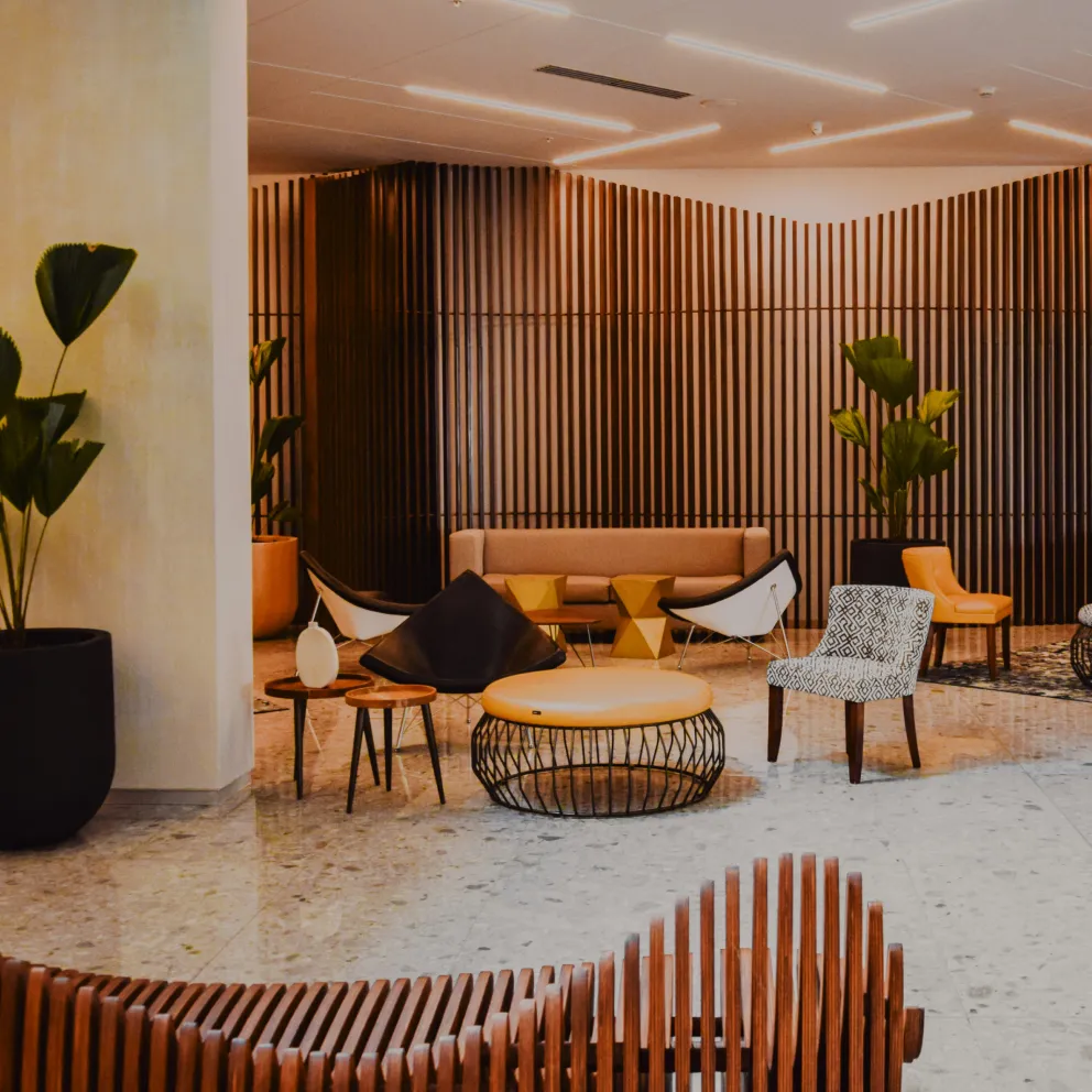 Modern lobby area with assorted chairs, circular ottomans, side tables, and large potted plants against a wooden slat wall.