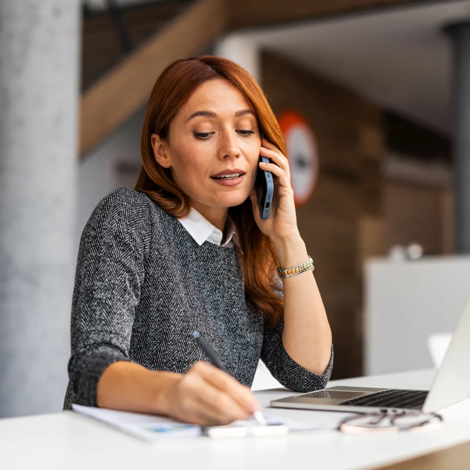 Woman talking on a smartphone while writing notes at a desk with a laptop and eyeglasses.