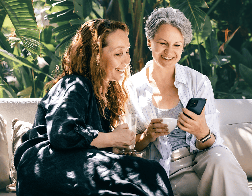 Two women sitting on a couch outdoors, smiling and looking at a smartphone while holding drinks.