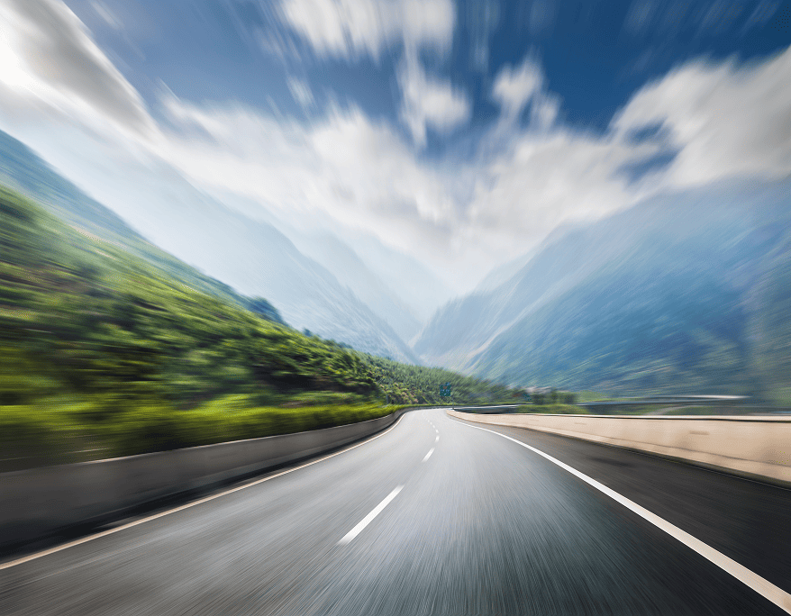 Curved empty highway with motion blur, surrounded by green hills and distant mountains under a partly cloudy sky.
