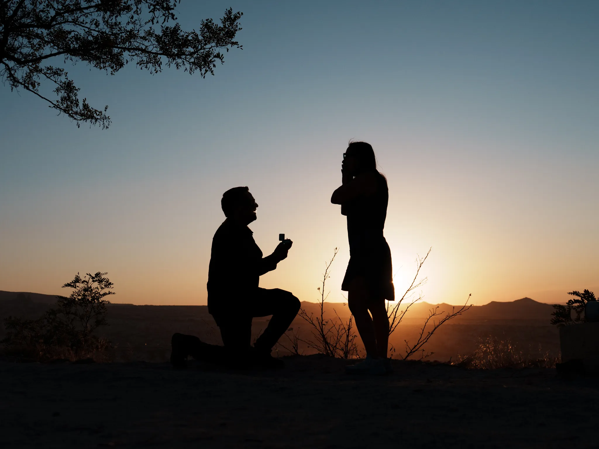 Documentary wedding photographer silhouette of a man proposing on one knee to his partner at sunset in the mountains