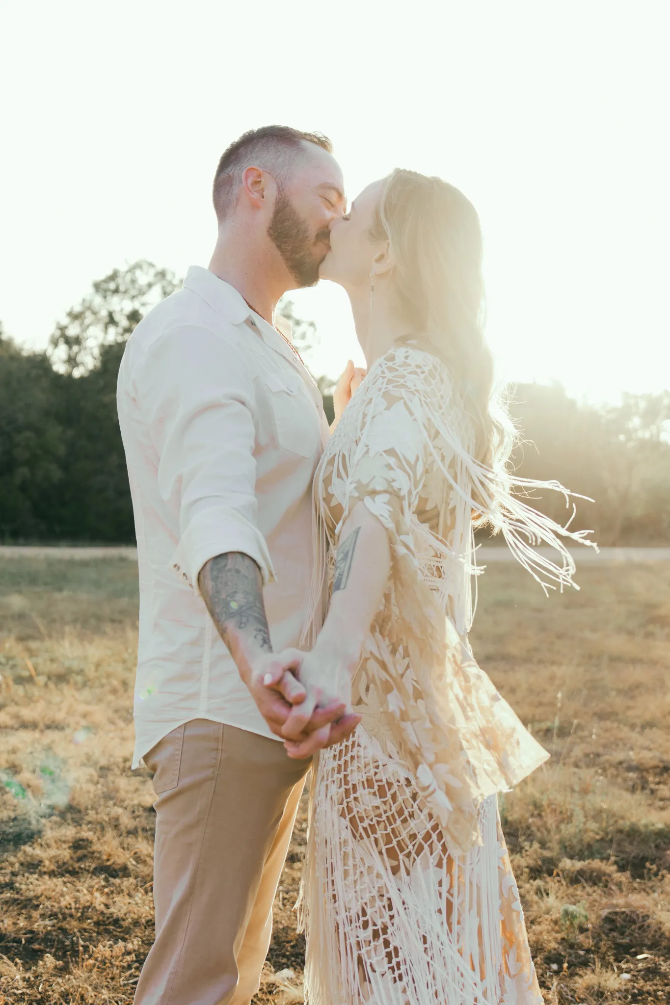 Documentary wedding photographer candid moment of a couple sharing a kiss at golden hour in an open field, bride in a bohemian fringe dress