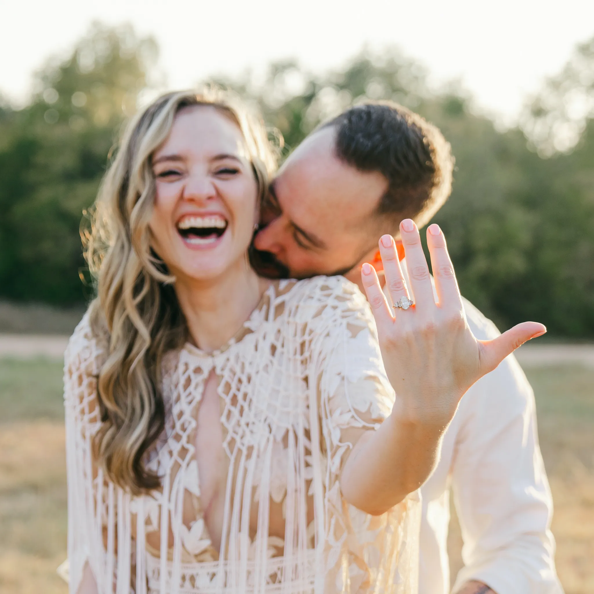 Documentary wedding photographer laughing bride showing off engagement ring while groom kisses her cheek at golden hour