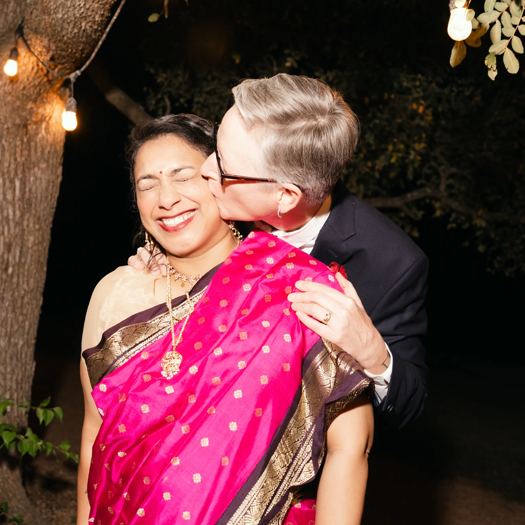 Austin Texas wedding photographer candid moment of two brides laughing under string lights at night one wearing a pink sari