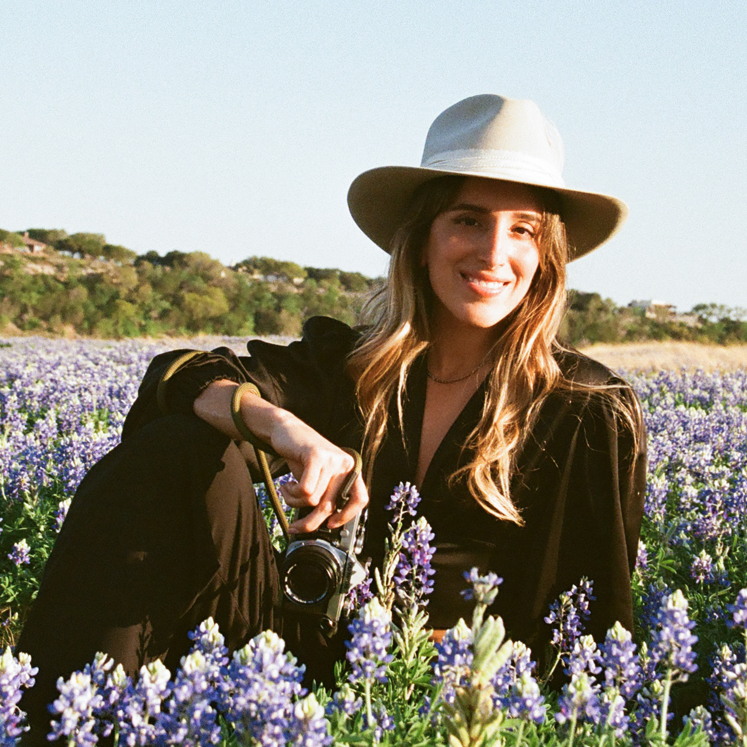 Austin wedding photographer Marcela Briere sitting in a field of bluebonnets, holding a film camera, wearing a wide-brim hat — Texas Hill Country