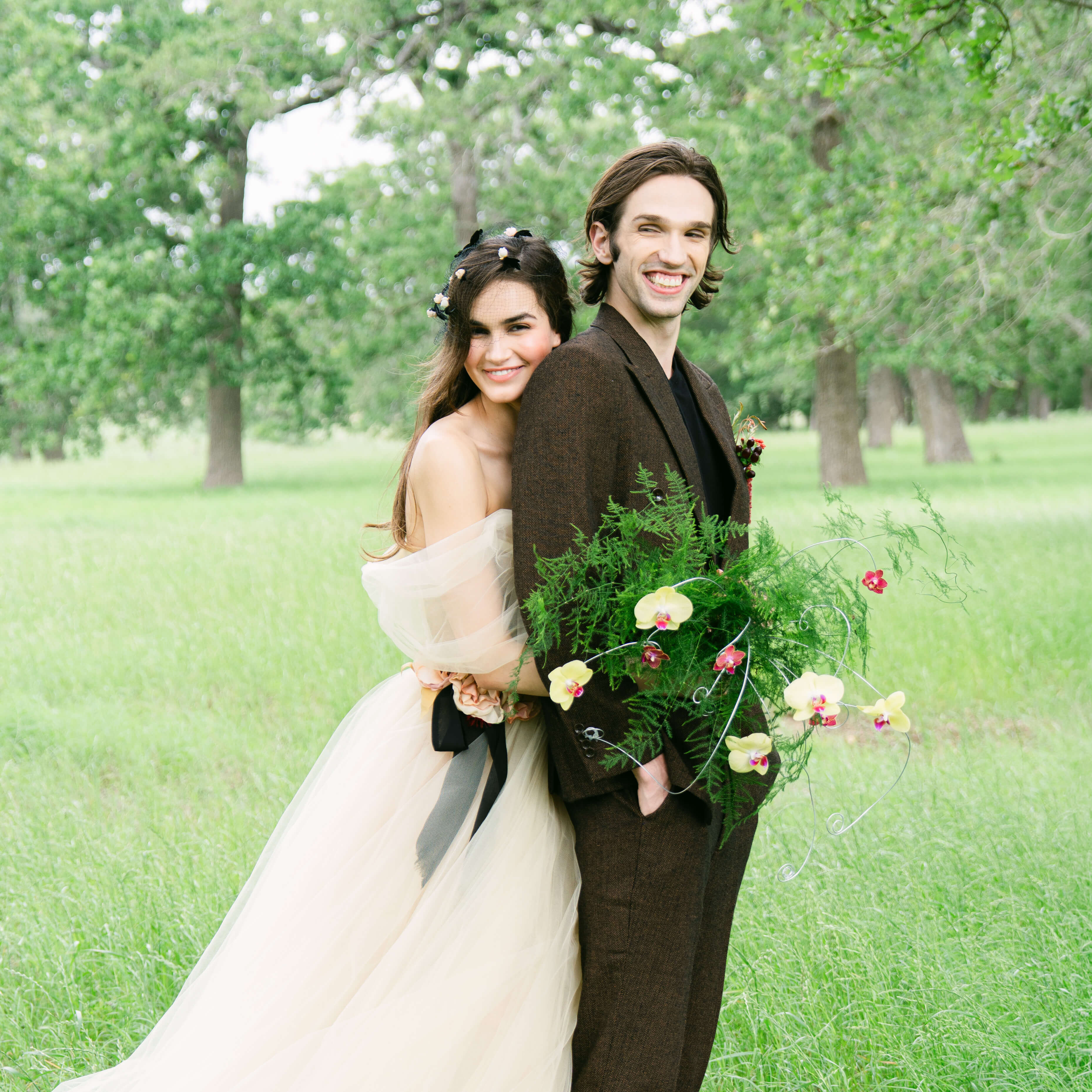 Austin Texas wedding photographer — bride in off-shoulder tulle gown and groom in brown suit holding bouquet of orchids and ferns in green Hill Country field