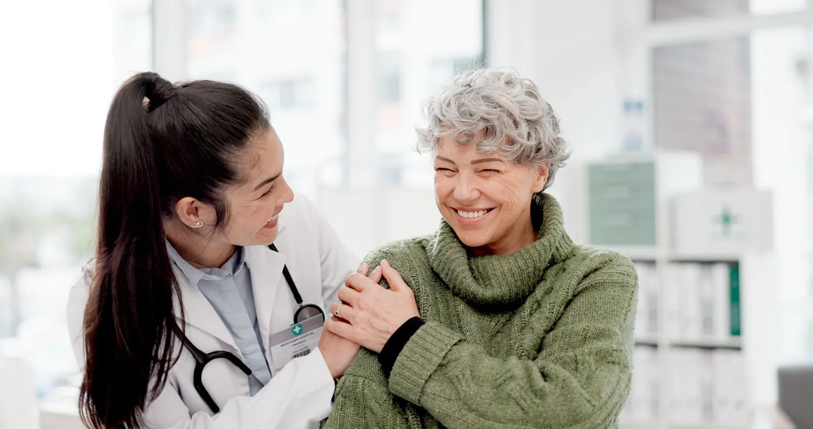 Smiling doctor with a stethoscope reassuring an elderly woman in a green sweater in a bright medical office.