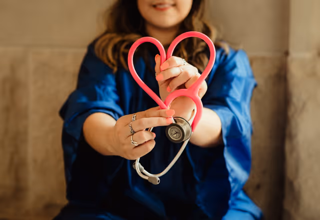A person in blue clothing holding a pink stethoscope shaped into a heart.