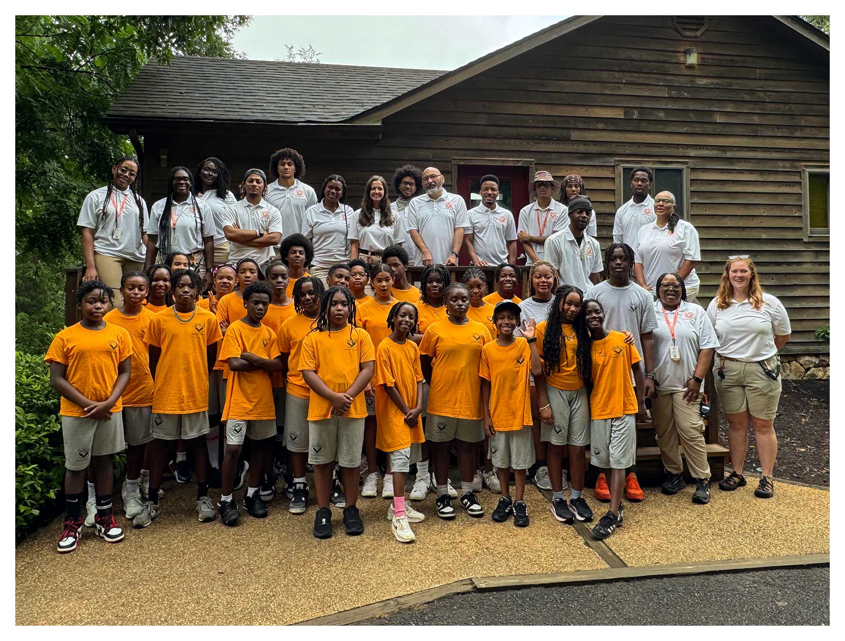 Group photo of campers at Camp Dogwood in orange shirts and beige shorts standing in front of a wooden cabin with adults in white shirts behind them.