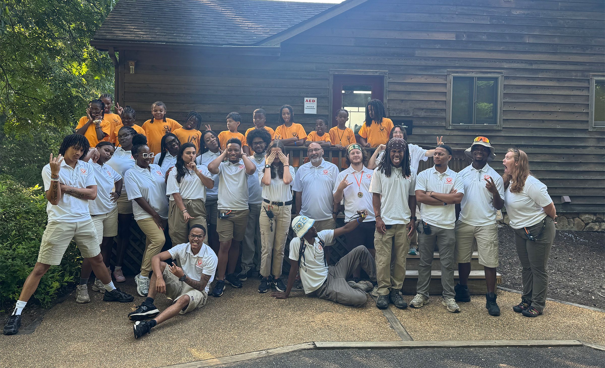 Group photo of diverse youth and adult staff at Camp Dogwood smiling and joking outside a wooden cabin at a summer camp.
